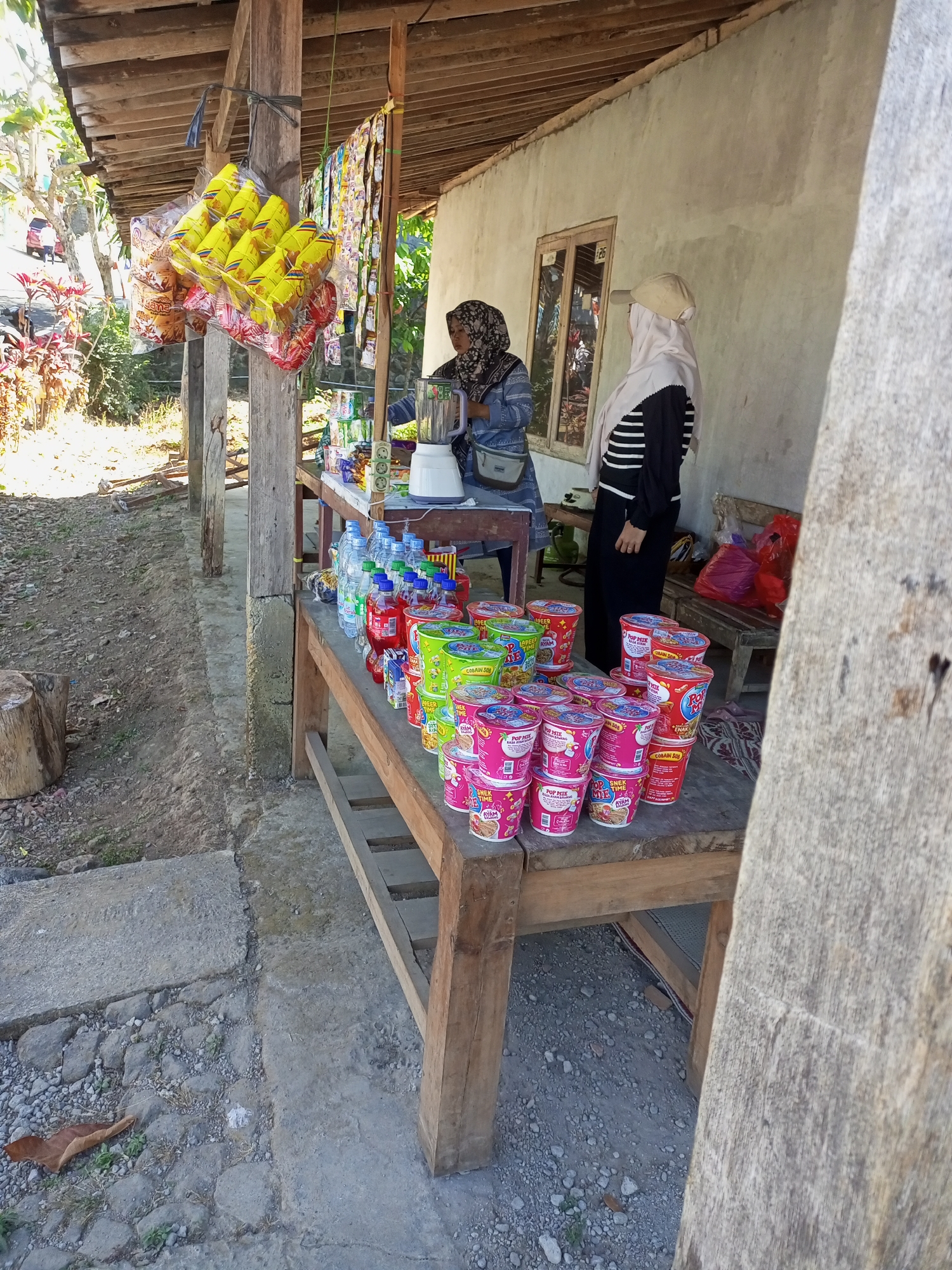 The image depicts an outdoor market stall in Jatipurno, Indonesia. Two women are present. One woman, wearing a headscarf and a blue patterned garment with a grey shoulder bag, is standing behind a counter where a white blender is visible. The second woman, also in a headscarf and a black and white striped shirt, stands to the right, facing away from the camera.
The stall is stocked with various items, including packaged snacks hanging from a wooden post, bottles of water and other beverages, and numerous colorful cups of instant noodles arranged on wooden tables. The stall has a rustic wooden roof supported by pillars. The ground is a mixture of dirt and concrete. In the background, a concrete wall with a window and some vegetation are visible. A red car can be seen in the distance on a road. The scene suggests a typical day at a local marketplace in a rural or semi-urban Indonesian setting.