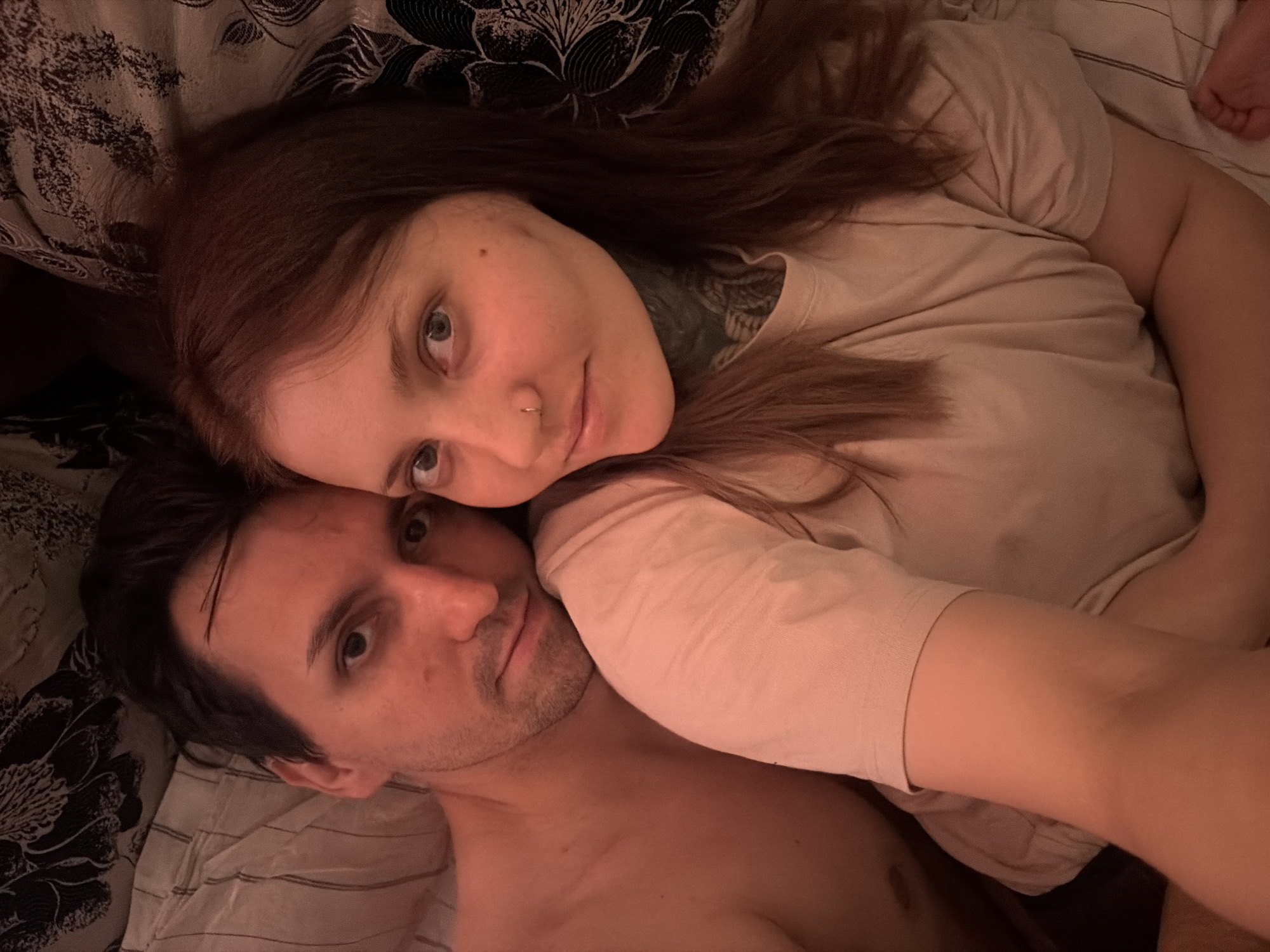 This is a close-up, overhead selfie of a couple lying in bed. The man, on the left, is partially visible, with his head turned towards the camera. He has dark hair and a light complexion. The woman, on the right, is more prominently featured, with her head resting on the man's shoulder. She has long, brown hair, light blue eyes, and a nose ring. She is wearing a light-colored t-shirt, and the man is shirtless. They are lying on a bed with striped white and grey sheets and a dark floral patterned duvet or headboard. The lighting is dim and warm, suggesting it may be nighttime or early morning. The overall impression is intimate and relaxed. The location is not specified in the image or its metadata.