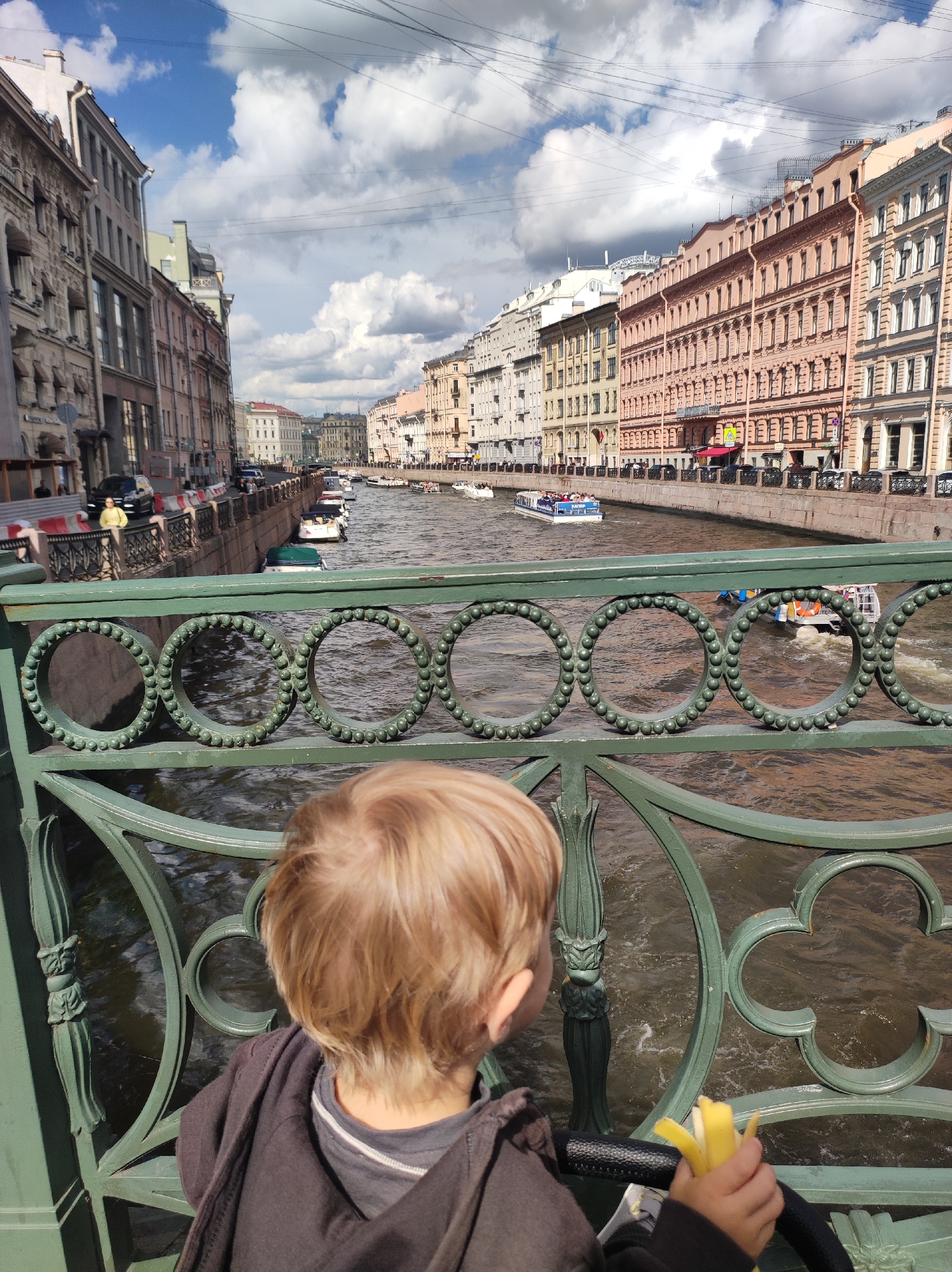 An over-the-shoulder medium shot captures a young child with blonde hair standing on a bridge in Saint Petersburg, Russia, looking out at a canal. The child, facing away from the camera, wears a dark grey hooded jacket over a grey shirt. The child's right hand is holding a peeled banana. In the background, the canal is lined with tall, colorful buildings in various shades of peach, pink, and beige, with ornate architectural details. Several boats are visible on the canal, including a larger passenger boat filled with people. The sky above is a mix of blue and white clouds. The foreground features a green decorative railing of the bridge, with circular patterns and floral motifs. The water in the canal appears brown and choppy, indicating movement from the boats and possibly wind. The overall scene suggests a sunny day in a historic urban setting.
