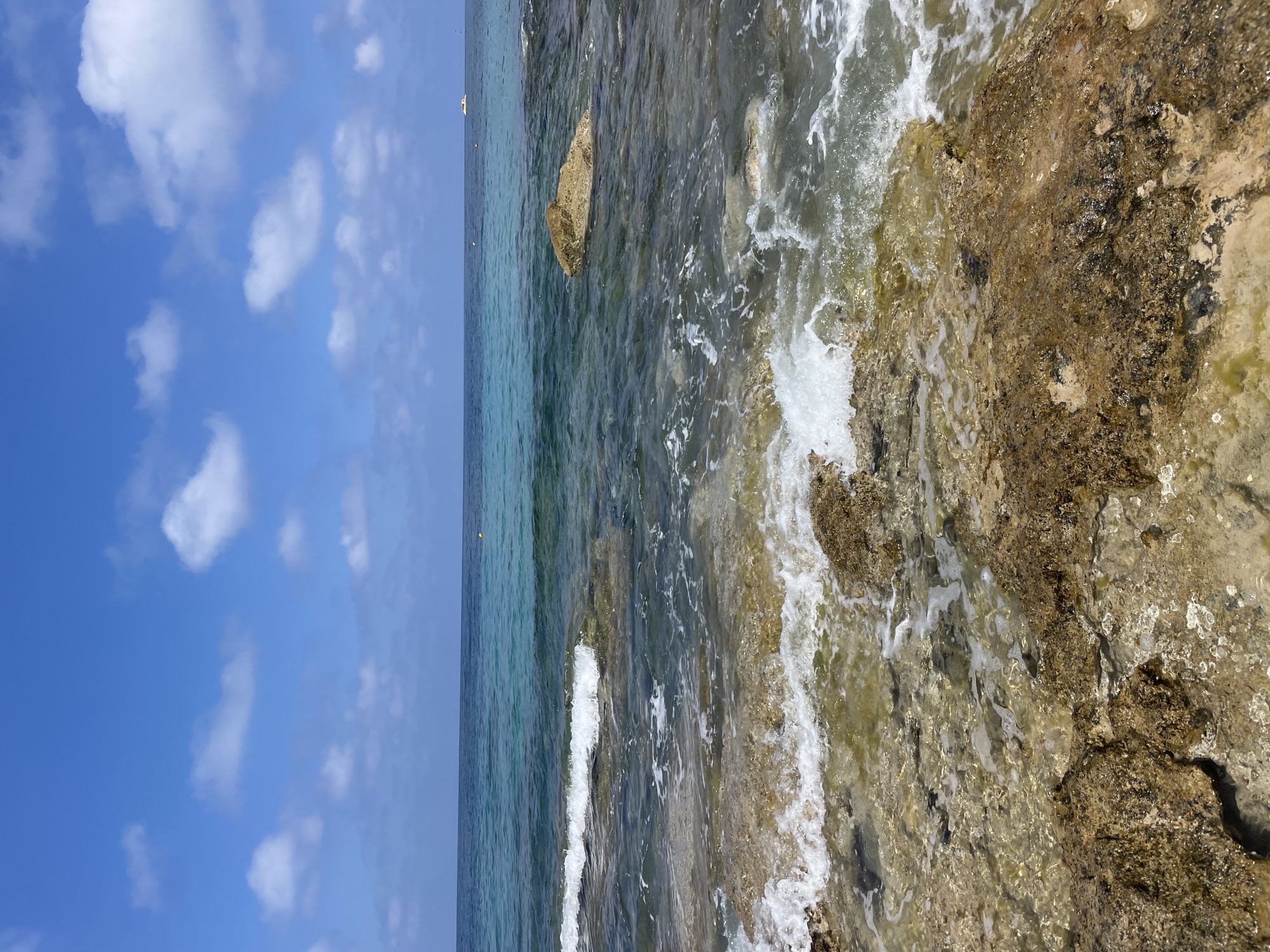 The image depicts a rocky coastline in the city of Paphos Municipality, Cyprus. The foreground is dominated by rough, wet rocks with some sea foam and small waves washing over them. The water is clear, revealing submerged rocks and a greenish hue in the shallow areas. Further out, the sea transitions to a deeper turquoise and then a dark blue as it meets the horizon. The sky is a bright blue with scattered white clouds. In the distance, a small white boat is visible on the water, and a yellow buoy floats to the left of the boat. The scene is natural and serene, with no people present.