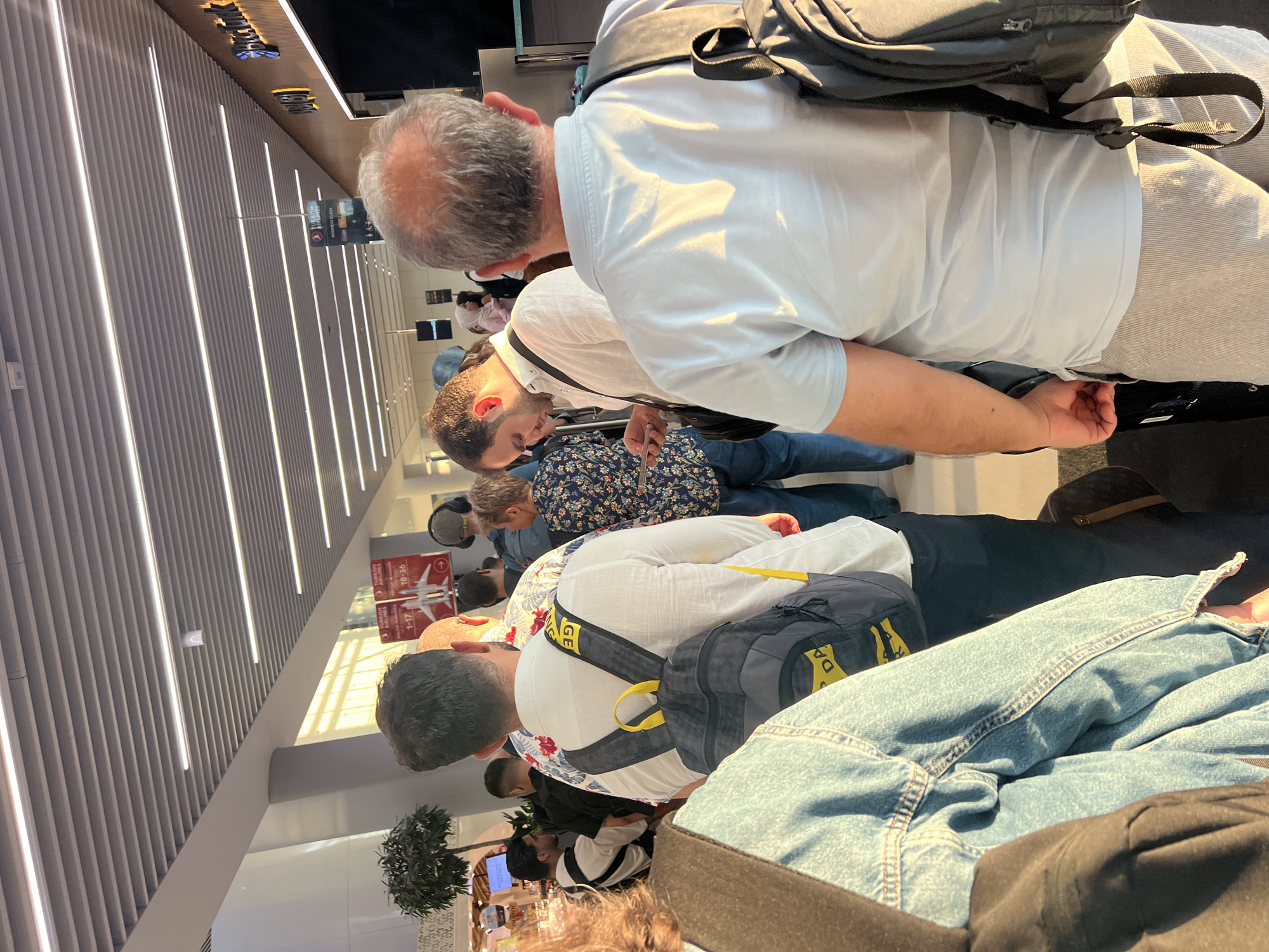 In Arnavutköy, Turkey, at an airport, individuals are standing in a queue. A person in the foreground wears a light blue polo shirt and light-colored shorts, carrying a backpack. To their left, a person in a denim jacket is visible. Behind them, a man in a white shirt with a floral pattern and a black backpack stands, looking down at his phone. Further back, a woman in a patterned blouse is also in the queue. The environment features modern airport architecture with a slatted ceiling and bright lighting. Signs for "Turkish Airlines" and flight information are present, indicating this is likely a check-in or boarding area.