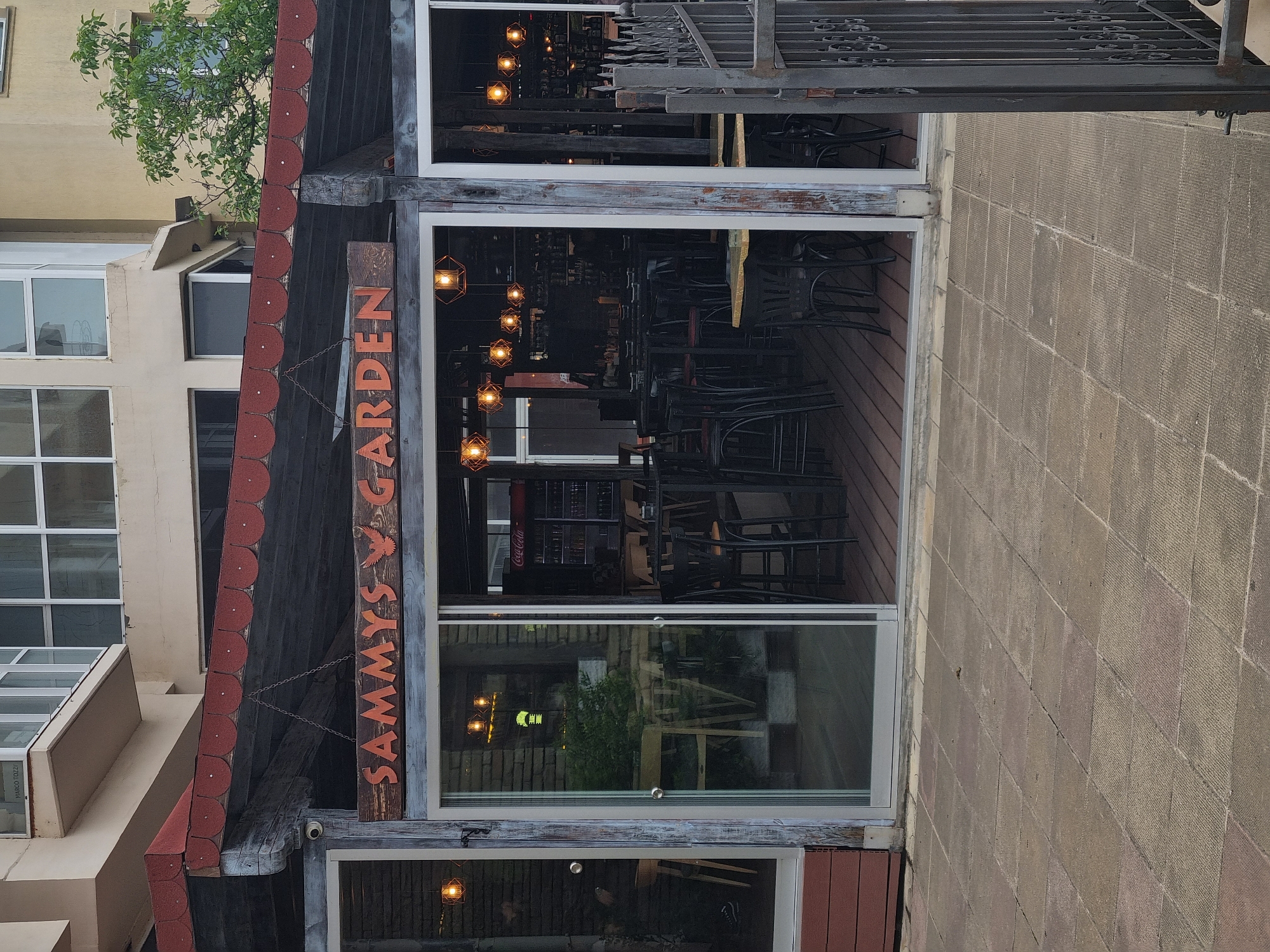 The image depicts the exterior of Sammy's Garden, a restaurant located in Veliko Tarnovo, Bulgaria. The restaurant's facade features a wooden sign with the name "SAMMY'S GARDEN" vertically oriented. A decorative roof with red trim covers the entrance. The restaurant is encased by a glass facade, which provides a view into the interior. Inside, multiple tables and chairs are visible, along with light fixtures hanging from the ceiling. In the middle pane of glass, a Coca-Cola refrigerator can be seen. The area outside the restaurant features a paved walkway, and a portion of an adjacent building can be seen, as well as metal decorative railings.