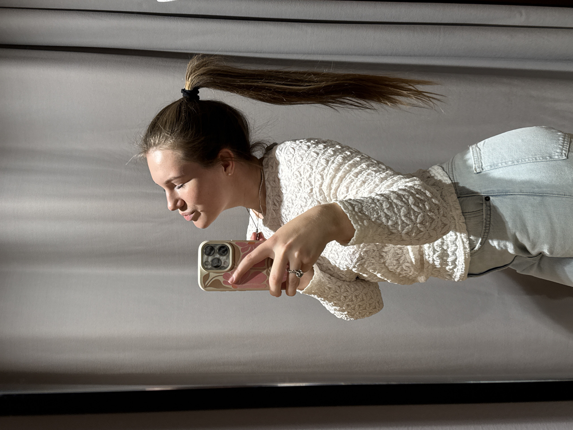 A young woman with a ponytail is holding a smartphone and taking a picture. She is wearing a white lace top and light blue jeans. The background appears to be a grey wall or curtain. The location cannot be determined from the provided image.