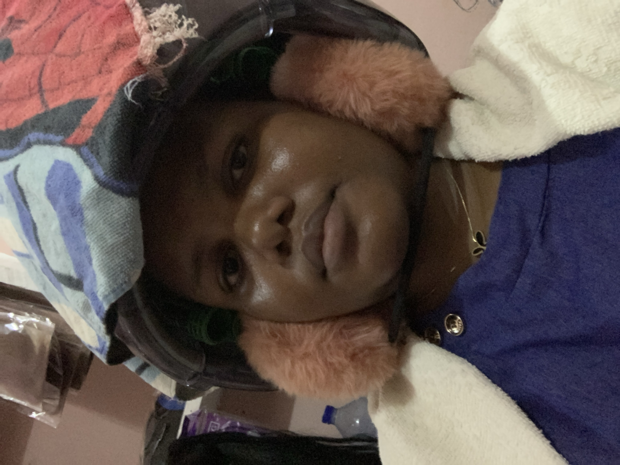 A person with dark skin is wearing a black helmet with furry pink ear covers, along with a blue shirt, a white towel, and a patterned towel. The location is not discernible from the image.