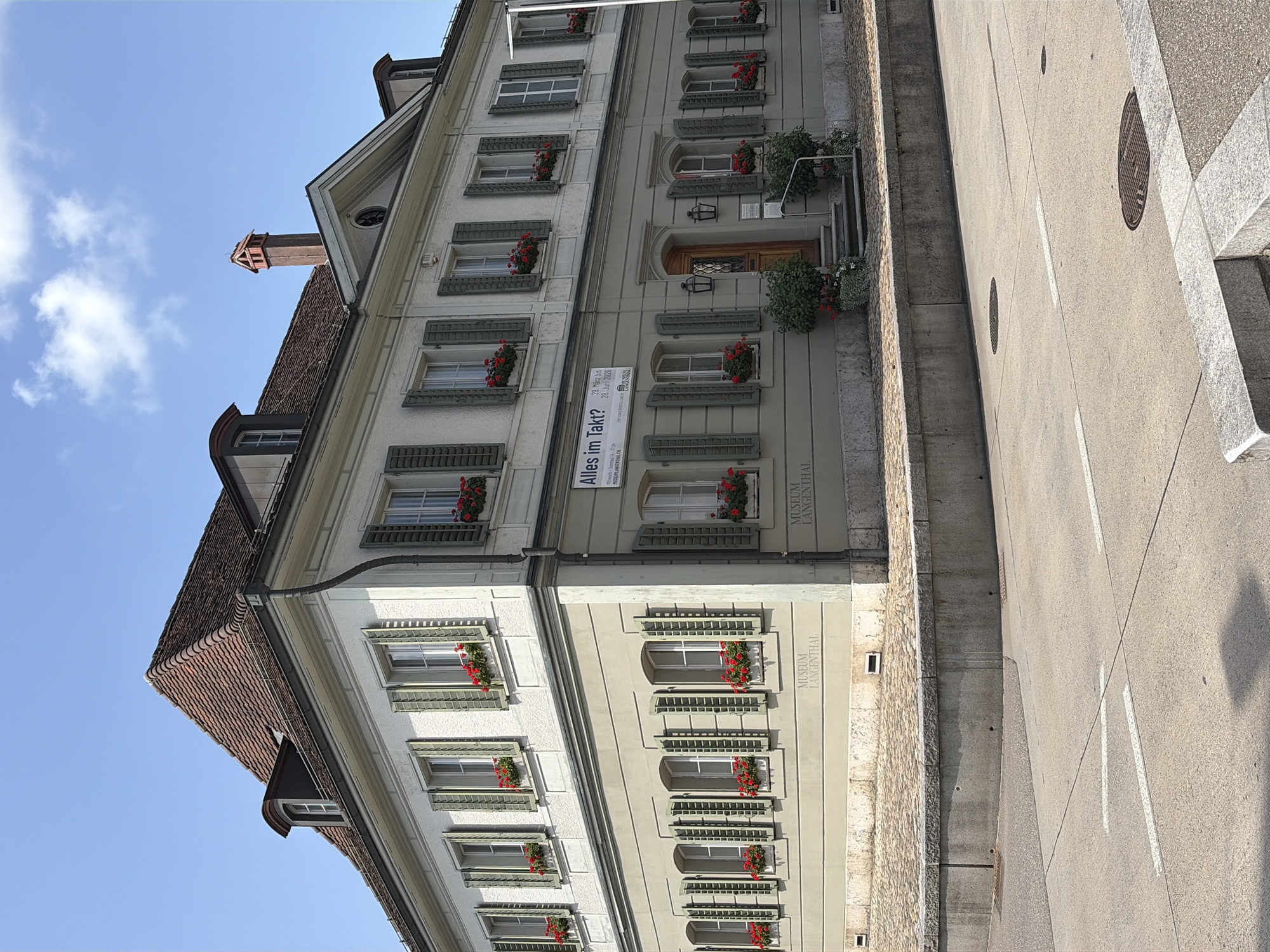 The image shows the Museum Langenthal, a two-story building with a light gray facade and a red-tiled roof. The building has numerous windows, most of which are covered by green shutters and adorned with red flowers in window boxes. The museum's name is visible in raised lettering on the lower part of the facade. A sign with the title "Alles im Takt?" is displayed on the upper floor, indicating an exhibition. The building is situated in a town square with cobblestone paving and a concrete retaining wall. A street with white lane markings and a manhole cover is in the foreground. The sky is clear blue with a few scattered clouds.