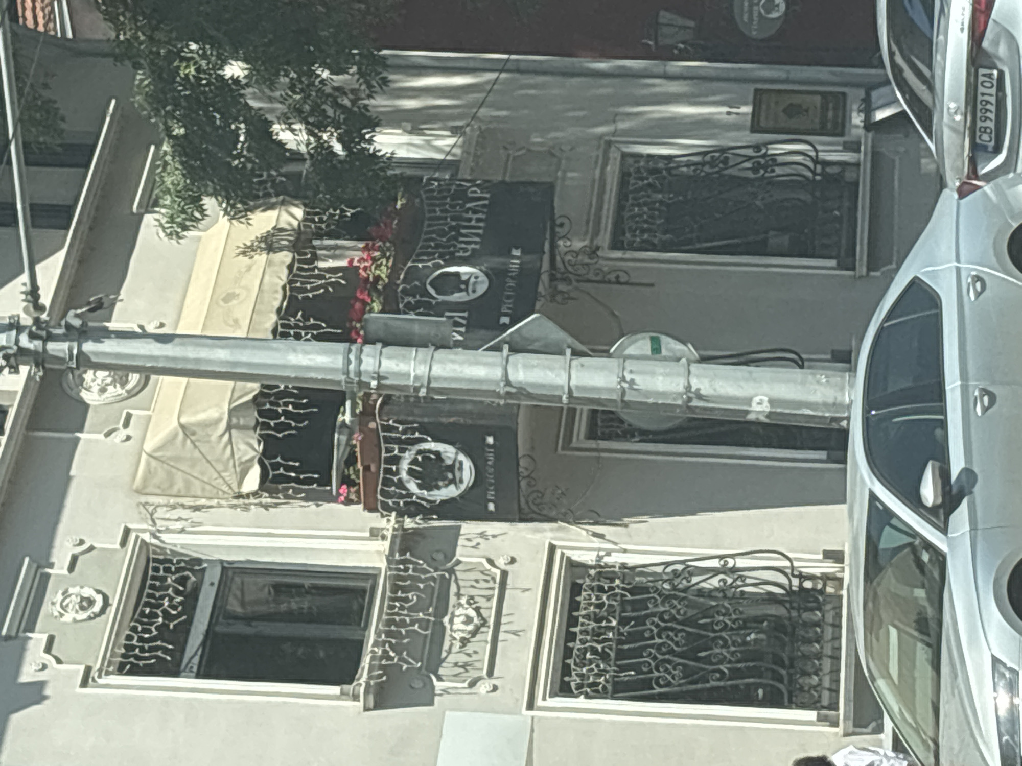 The image shows the exterior of a building in Sofia, Bulgaria, with a street and cars in the foreground. The building has a light-colored facade with decorative elements around the windows. There is a balcony on the second floor with planters containing red flowers and a black sign that reads "РЕСТОРАНТ ЧИНАР" (Restaurant Chinar). A metal pole, likely a street light or utility pole, stands in front of the building. Two silver cars are parked on the street, one of which has a license plate that reads "CB 9991 OA". A person in a white shirt is partially visible on the left side of the frame, standing near a window. Trees with green foliage are visible on the right side of the image. The scene appears to be a daytime shot on a sunny day.