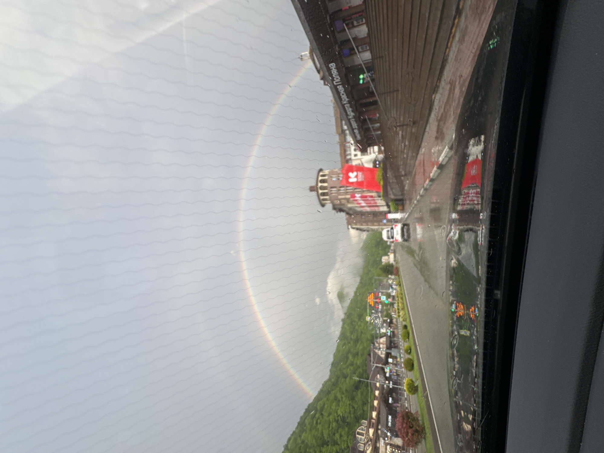 The photograph, taken from inside a vehicle, captures a view of Estosadok, Russia, under rainy conditions. A full rainbow arcs across the sky, extending from the left side of the frame over the town. The foreground consists of a rain-streaked vehicle window.
The middle ground presents a street scene, reflecting the rainy environment. Buildings of varying heights, including some with red signage, line the street. A white vehicle is present on the road.
The background displays a forested hillside, providing a natural contrast to the urban elements. The sky is overcast, with grey clouds dominating the upper part of the frame.