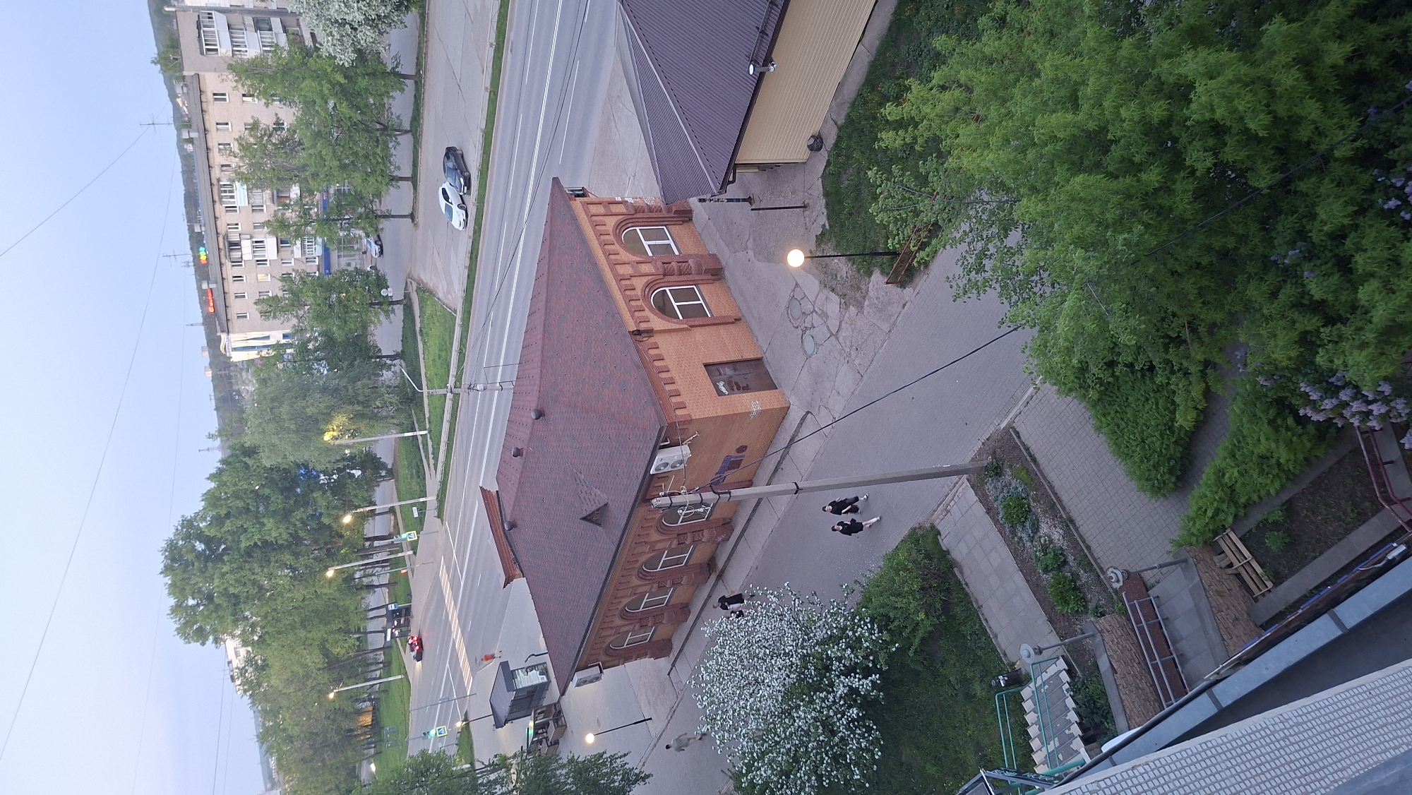 The image shows an aerial view of a section of the city of Chaikovsky, Russia. In the foreground is a building with a distinctive red-brown roof, beige-brick facade, and arched windows. Two people are walking on the sidewalk adjacent to the building. A street runs parallel to the building, with cars visible on the road. Rows of trees line the street, and lamp posts illuminate the sidewalk. A multi-story apartment building is visible in the background. The environment appears to be a mix of residential and commercial areas.
