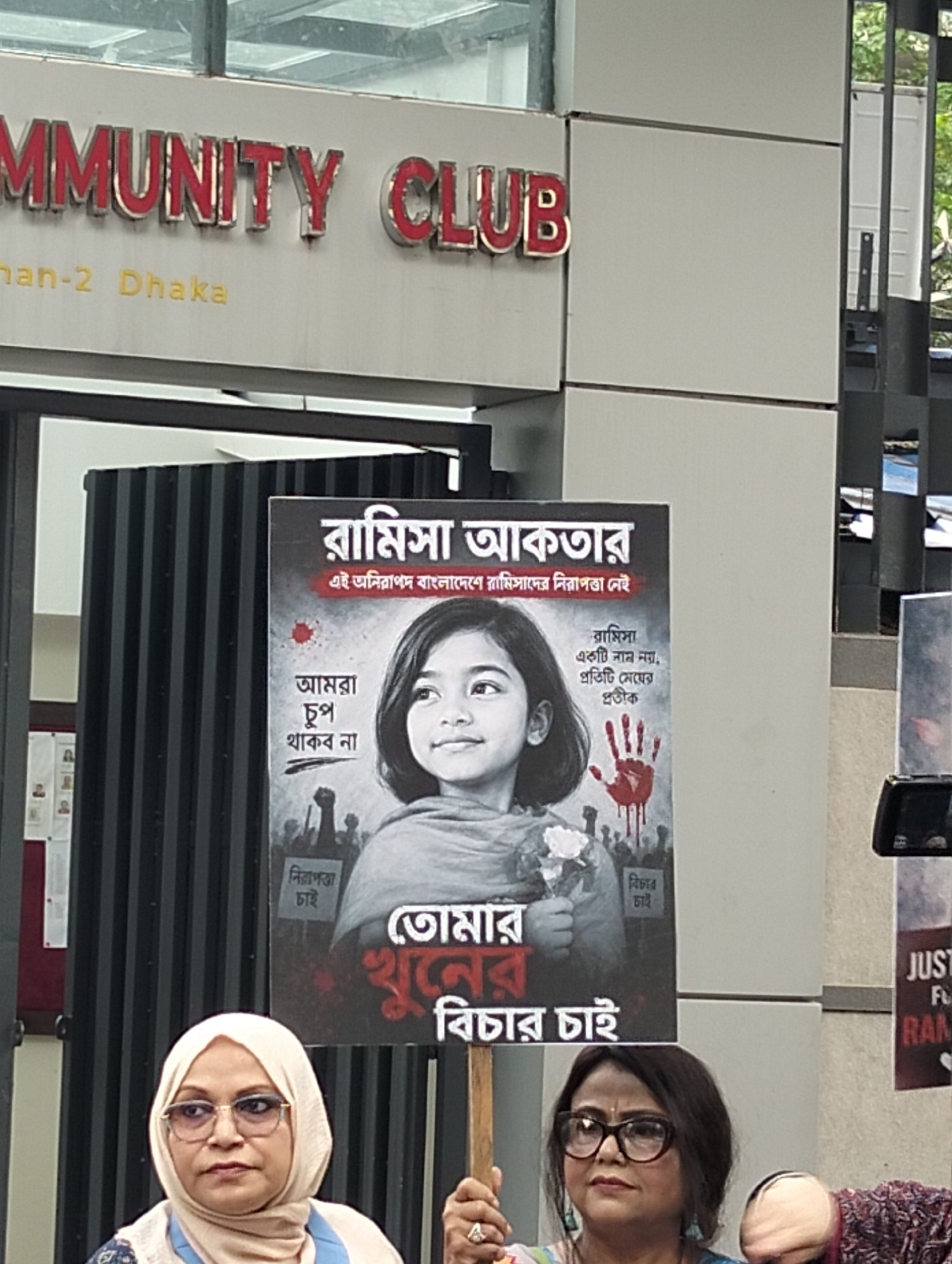 The image captures a scene at the Community Club in Dhanmondi-2, Dhaka. A sign above the entrance of the club, reads "MMUNITY CLUB, Dhanmondi-2 Dhaka". In the foreground, two women hold a poster bearing an image of a young girl named "Ramisa Aktar" and a caption that reads, "We will not be silent. We want safety. Justice for your murder." The poster seems to be part of a protest or campaign, possibly related to child safety or justice. The background includes architectural details of the Community Club and hints of greenery.