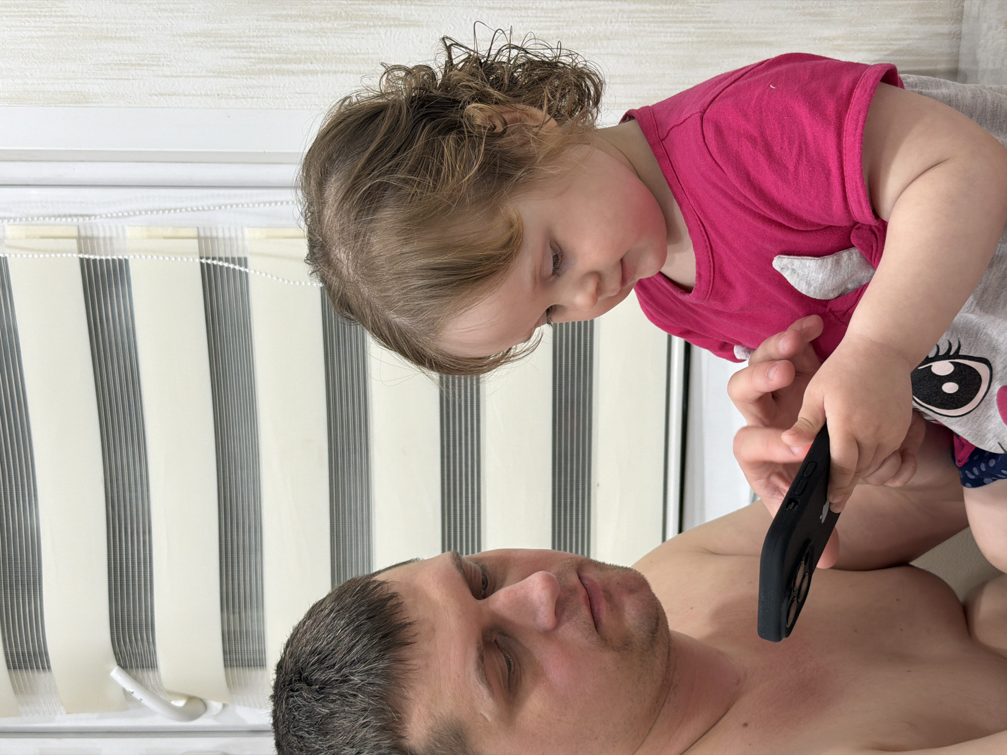 A toddler is using a smartphone to take a picture of a man lying down. The child, wearing a pink shirt and a gray printed top, is holding the black smartphone. The man has short, dark hair and appears to be relaxed. The location is Chaikovsky, Russia. The background includes vertical blinds and a wall with a textured surface.