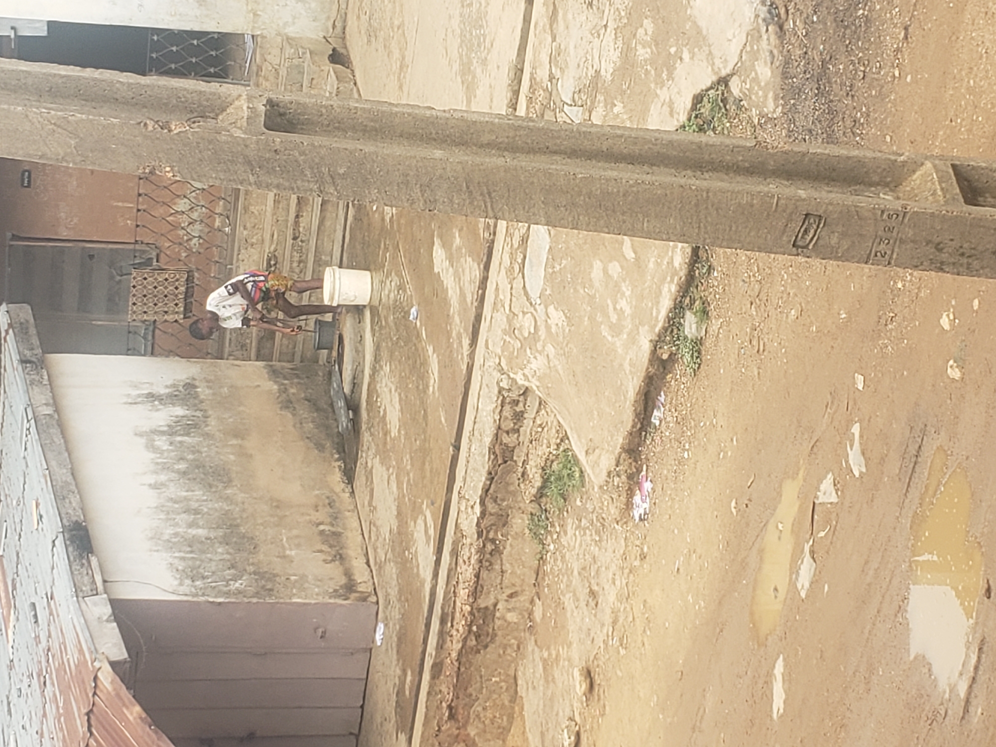 The image depicts a scene in Alalubosa, Nigeria. A person stands near a building, interacting with a bucket. A long concrete structure is visible overhead. The surrounding area appears to be an unpaved street with puddles.