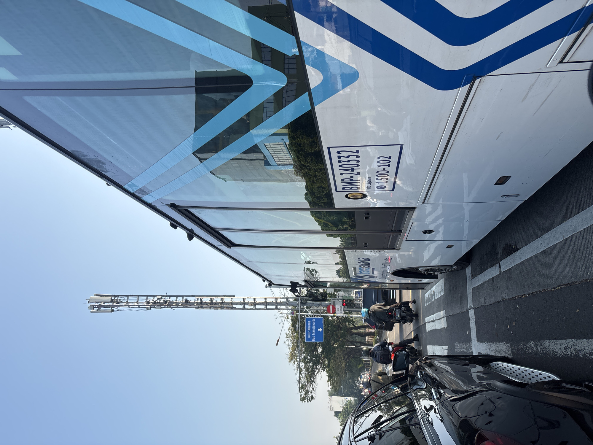 In West Jakarta, Indonesia, a bus transit lane is depicted in a traffic scene under a clear sky. A large white Transjakarta bus, with blue V-shaped accents and text indicating "BMP-240332" and "1500-102", is positioned on the right side of the frame, facing away from the camera. A black SUV is visible to the left, parked partially in the frame, showcasing its side mirror and running board. Several motorcycles, with riders wearing helmets, are in motion on the road in front of the bus. A traffic sign in blue with white text reads "Jalur Khusus Bus Transjakarta" (Exclusive Bus Transjakarta Lane), with an arrow pointing downwards, suggesting the bus lane's direction. A tall cell tower stands prominently in the background, with trees and a building also visible. The ground is paved, with white crosswalk markings indicating a pedestrian crossing.