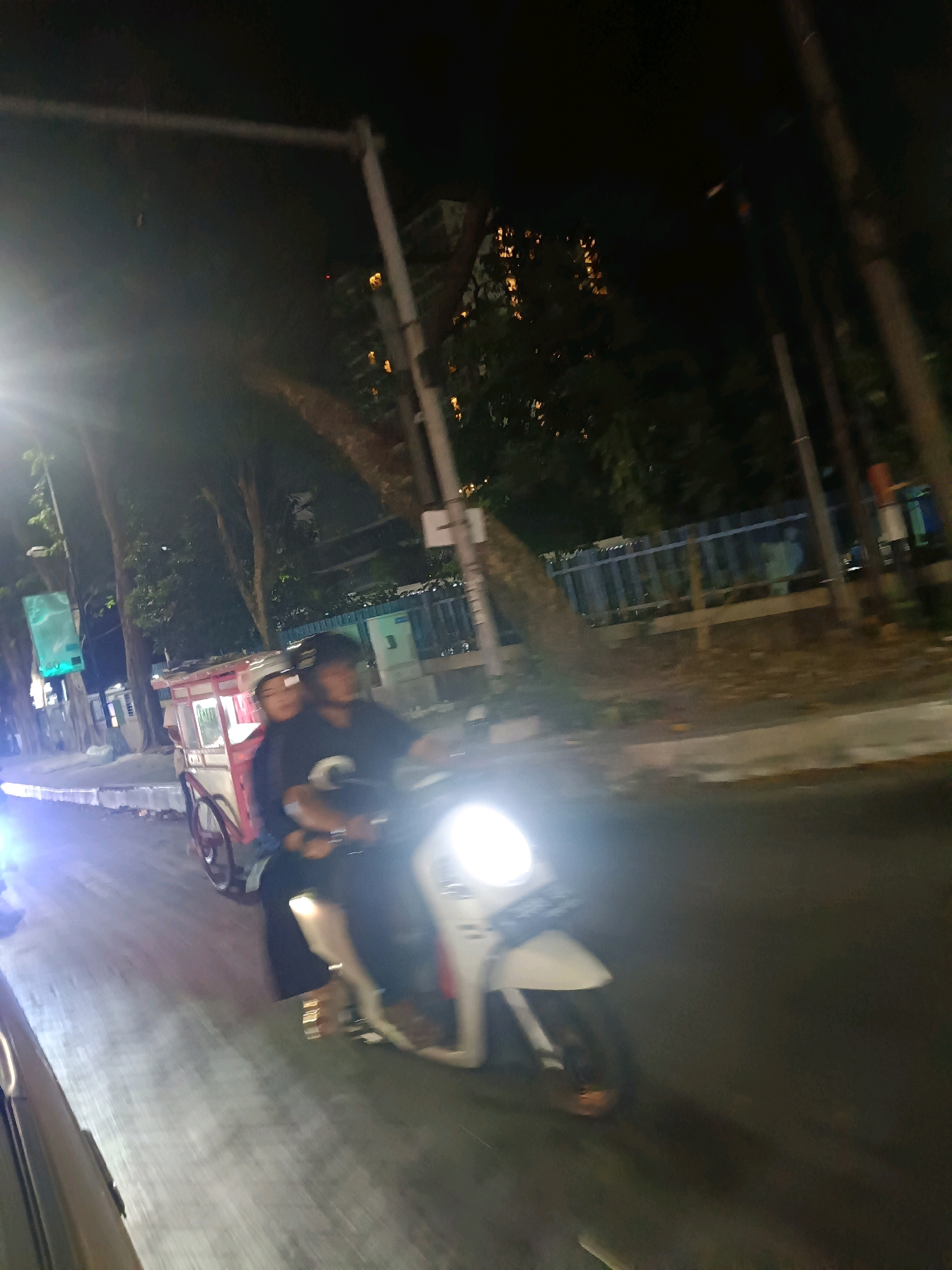 The image depicts a nighttime street scene in Surabaya, Indonesia, featuring two individuals on a motorcycle. The driver, wearing a dark shirt and helmet, is operating the motorcycle, while a passenger, also wearing a dark shirt and a cap, is seated behind them. A red and white food cart is visible next to the motorcycle. The street is paved and appears to be illuminated by streetlights and the motorcycle's headlight, which casts a strong glare. Trees and a fence line the side of the street. The overall image is somewhat blurry due to motion.