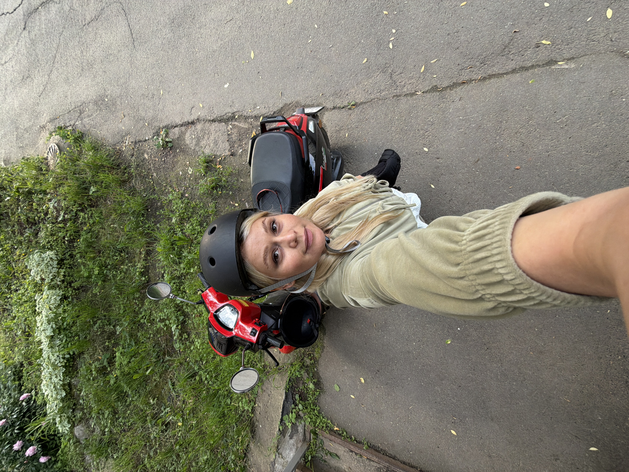 A woman with blonde hair and a black helmet is taking a selfie while standing next to a red and black scooter on a paved surface in Sarapul, Russia. She is wearing a light green hooded jacket and dark socks. The scooter is parked on the right side of the frame, with the front end pointing towards the top left. Behind the scooter is a grassy embankment with some white-leafed plants and pink flowers. The ground is asphalt with some scattered leaves.