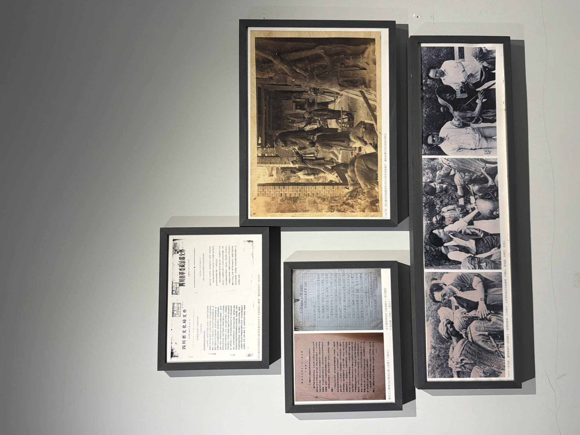 This image captures a gallery wall displaying five framed exhibits, comprising official documents and photographs, situated within the Chongqing High-tech Zone, China. The top-left frame contains two side-by-side Chinese documents. The left document is titled "四川省文化局文件" (Sichuan Provincial Cultural Bureau Document), dated 1959. The right document, dated 1978, is titled "四川省革委政治..." (Sichuan Provincial Revolutionary Committee Political...). A textual label below this frame clarifies these are "Documents from Sichuan Provincial Cultural Bureau and Sichuan Provincial Revolutionary Committee regarding the 'Rent Collection Courtyard' sculpture's 'reproduction and recreation'". Below this, on the middle row, the left frame holds two documents. The left document is a typed Chinese report titled "重庆工人雕塑活动情况汇报 (初稿)" (Report on Chongqing Workers' Sculpture Activities (Draft)). The right document is a handwritten Chinese discussion outline titled "关于复制再创作《收租院》一些问题的讨论提纲 (初稿)" (Discussion Outline on Some Issues Regarding the Reproduction and Recreation of "Rent Collection Courtyard" (Draft)). To the right, on the middle row, a large, sepia-toned photograph is displayed. It depicts an interior or semi-outdoor workshop scene from 1957. Multiple individuals, presumably sculptors or art students, are actively working on large, figurative sculptures, some of which appear to be human forms in various stages of completion. Scaffolding and tools are visible around the figures. A label beneath this frame states: "1957, Teachers and students from the Sculpture Department of Sichuan Academy of Fine Arts reproducing and copying Dazu Rock Carvings for the study of national heritage, on-site photo." The bottom row features a single, wide frame containing three distinct black-and-white photographs, chronologically arranged from left to right, taken between 1973 and 1978. * The left photograph shows a man in sunglasses and a short-sleeved shirt interacting with a group of sculptures depicting laborers carrying baskets, with other individuals observing. * The middle photograph shows a different man in a light-colored shirt and sunglasses standing beside a sculpture of an elderly person wearing a hat and holding a tool, with another person in a dark shirt nearby. * The right photograph depicts two men in light-colored shirts observing a sculpture of a figure next to a water buffalo, with one man gesturing towards the artwork. A label below this frame identifies these as "working photos of the reproduction and recreation of 'Rent Collection Courtyard' (Tangxi Foundry)," listing the individuals from left to right as Long Dehui, Ye Jinshan, Guo Xuanchang, Wu Mingwan, and Wang Yuyi. The exhibits are mounted on a plain, light-grey wall, illuminated
