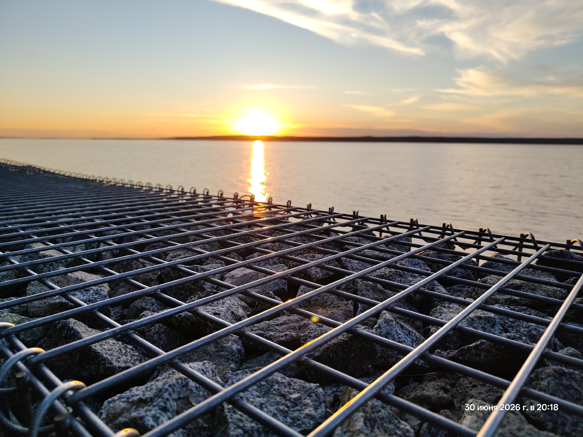 An outdoor, eye-level, medium shot captures a gabion wall in the foreground, composed of a metal mesh enclosing rocks. The wall runs diagonally from the lower left to the upper right of the frame. Beyond the gabion wall, a body of water stretches to the horizon. The sun is setting over the water, casting a bright, golden reflection on its surface. The sky above transitions from a pale blue at the top to hues of orange and yellow near the horizon, with wispy clouds present. A date and time stamp, "30 июня 2026 г. в 20:18", is visible in the lower right corner. The scene is devoid of people or animals.