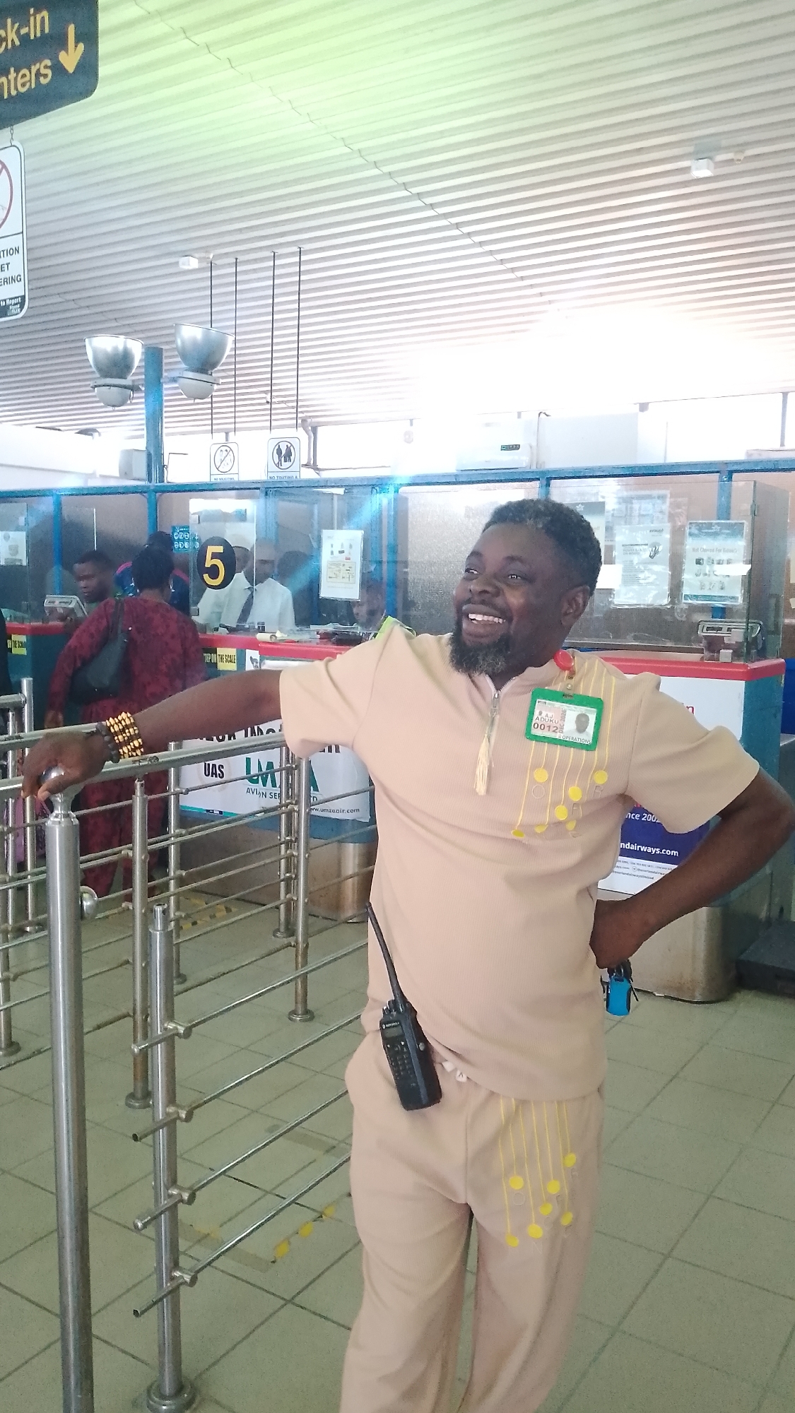 A man is standing inside what appears to be an airport check-in area. He is leaning against a silver metal railing, smiling and looking towards the right. He is wearing a light brown, short-sleeved tunic-style shirt and matching pants, both with yellow decorative accents. A green identification badge is pinned to his shirt. A black walkie-talkie is clipped to his pants. On his left wrist are multiple gold bracelets.
Behind the man are check-in counters with the number "5" displayed above one. People are standing in line or behind the counters. Signage is visible in the background, including a "Check-in" sign with a downward arrow.
The scene appears to be indoors, in a brightly lit, modern airport or travel hub with tiled flooring. The text on the badge reads "ADUKU 0012 OPERATIONS". The text visible on the advertising suggests the image was taken in Nigeria. Given the advertising for "Umzaair", which is based in Orerokpe, Nigeria, the image was likely taken there.