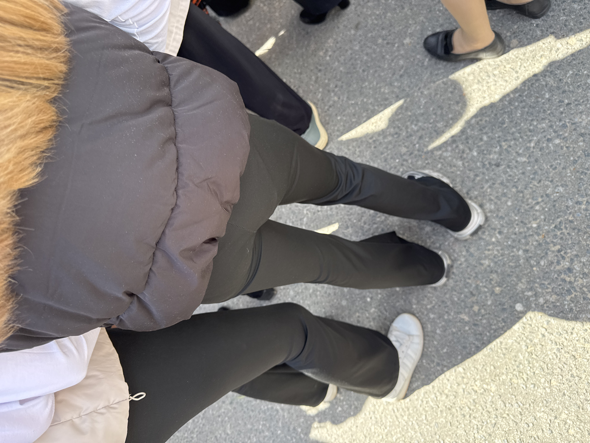 The image shows a view of legs and shoes on an asphalt surface. Several individuals are present, wearing dark pants and a variety of shoes including sneakers and flats. A partial view of a puffy jacket with fur trim is also visible. The location appears to be an outdoor urban environment with shadows indicating sunlight. The image does not contain information to determine if the location is Yamyshevo, Russia.