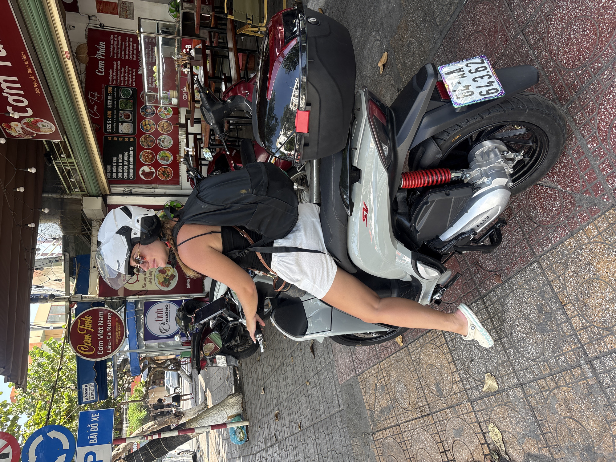 A person is captured in a playful or relaxed pose, leaning on a light grey scooter on a sunny day in what appears to be a city in Vietnam. The person wears a white helmet, sunglasses, a black tank top, white shorts, and white Crocs. The scooter has a black storage box attached to the rear and features a red shock absorber. The visible license plate reads "58A1 - 643.62". The setting is a bustling street with a patterned tiled sidewalk, lined with various storefronts, indicative of a commercial area. Bright sunlight suggests it is daytime with clear weather.
Prominent in the background are several restaurant signs with Vietnamese text. One large sign advertises "Cơm Phần" (rice set meals) with a detailed menu showing various "Cơm" dishes like "Cơm sườn" and "Cơm gà", along with prices (e.g., "SIZE S: 40K", "SIZE M: 50K"). Another sign for "Cơm Tấm" is visible. A circular sign for "Cơm Tinh - Cơm Việt Nam - Lẩu Cá Nướng" displays the address "205 Nguyễn Bỉnh Khiêm" and a phone number "0935 926 911". Other signs include "Tình NƯỚNG" and "Bãi Đỗ Xe" (Parking lot). Trees and other buildings can be seen further down the street.