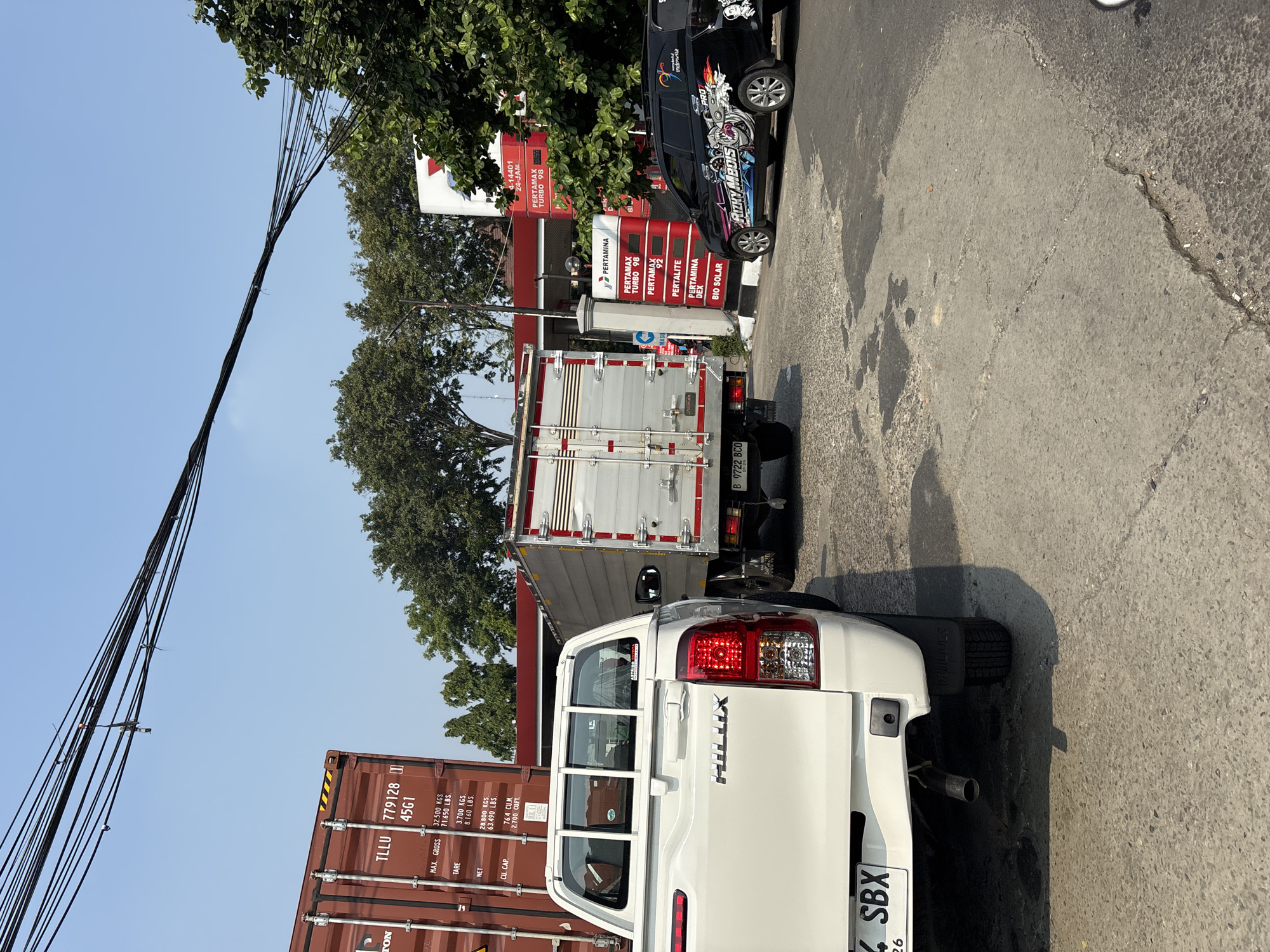 The image captures a daytime scene at a Pertamina gas station in North Jakarta, Indonesia. In the foreground, the rear of a white Toyota Hilux pickup truck is visible, with its license plate reading "B 14 SBX". To the right of the pickup, a black SUV is parked, adorned with a colorful decal. Behind the pickup, a silver box truck is lined up, its license plate reading "B 9722 BCO". Further back, a large red shipping container with the marking "TLLU 779128 45G1" is visible. A red and white Pertamina gas station structure with pumps and signage displaying fuel prices like "PERTAMAX TURBO 98", "PERTAMAX 92", "PERTALITE", "PERTAMINA DEX", and "BIO SOLAR" is situated in the background. Overhead, a network of dark electrical wires crisscrosses the clear blue sky. The ground is paved with concrete, showing signs of wear and tear in places. The scene suggests a typical day at a busy urban gas station.