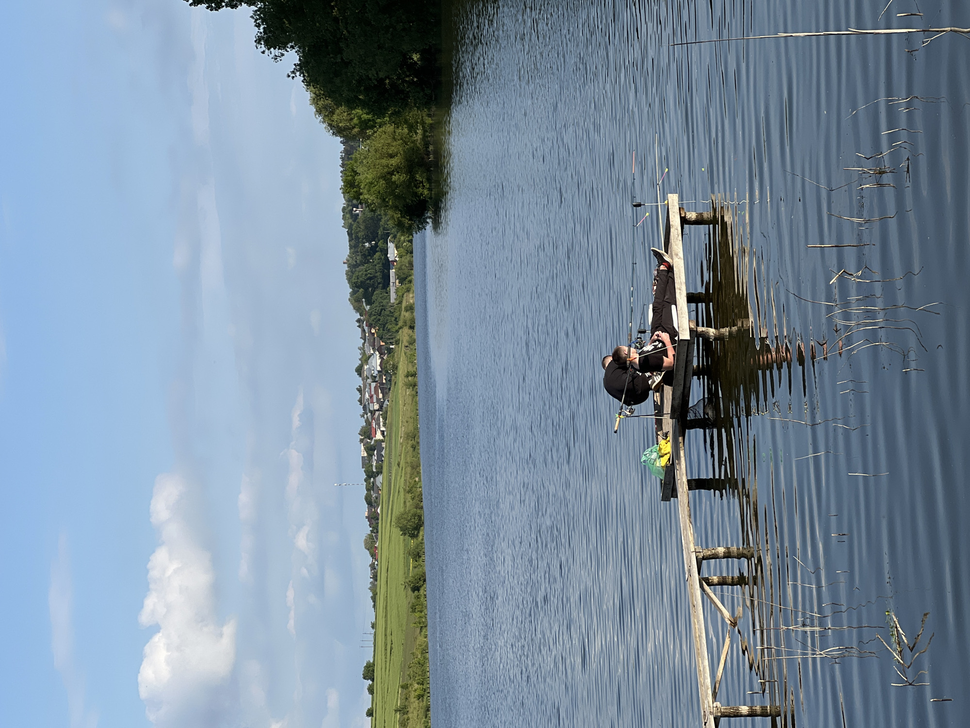The image captures a peaceful daytime scene of two individuals fishing on a wooden pier in Bazaryntsi, Ukraine. The pier extends into a calm body of water, likely a lake or wide river, which occupies the middle ground of the image. The water surface shows gentle ripples, reflecting the clear sky.
On the pier, one person, dressed in dark clothing, is seated and engaged in fishing, with multiple fishing rods set up. A second person is partially visible behind them. A yellow or light green bag is placed near their gear. The pier itself appears to be constructed from sturdy, rustic wooden beams and planks. Dried reeds and small pieces of natural debris are visible floating on the water near the pier.
The left side of the frame showcases a scenic landscape. A green shoreline with lush trees and fields stretches along the water's edge. In the distance, a small village or town with numerous houses and a prominent tall antenna or tower can be seen, nestled against rolling hills. The sky above is bright blue with scattered white, fluffy clouds, indicating fair weather. The overall ambiance is tranquil and serene, characteristic of a leisurely day spent fishing in nature. No text is visible in the image.