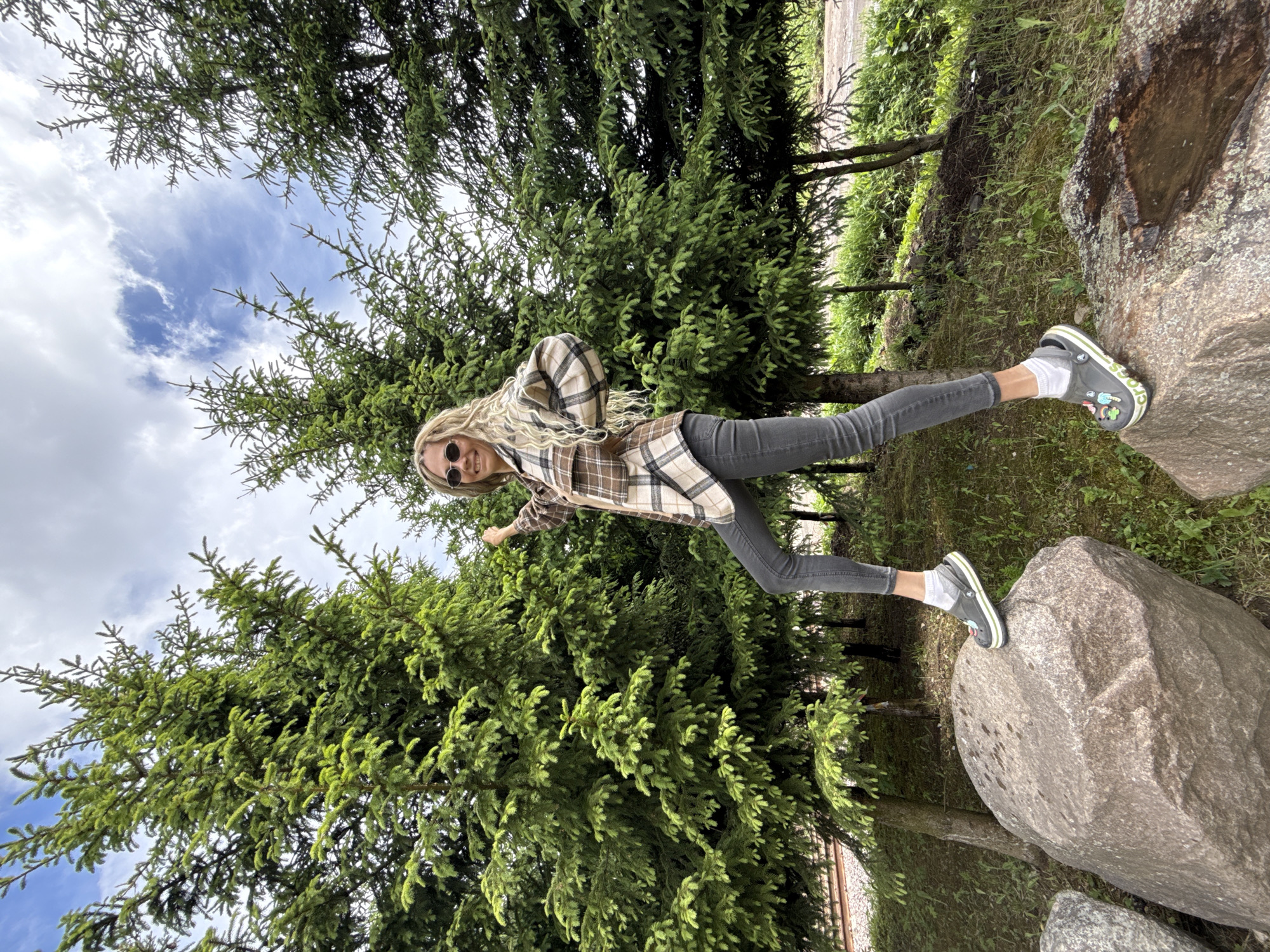A young woman stands with one foot on a boulder, her other foot balanced on a smaller rock in a wooded area. She wears jeans, a plaid shirt, sunglasses, and Crocs with socks. Coniferous trees surround her, and a patch of blue sky with white clouds is visible above. The location is Rusanovo, Russia.