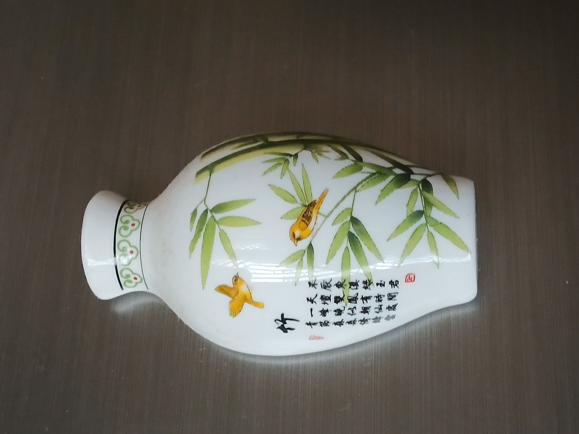 A white, rectangular vase with a cylindrical neck is lying on a metallic surface. The vase is decorated with images of bamboo stalks and two small yellow birds. Chinese calligraphy, in black ink, is inscribed on the side of the vase. The background is a neutral gray metallic surface.