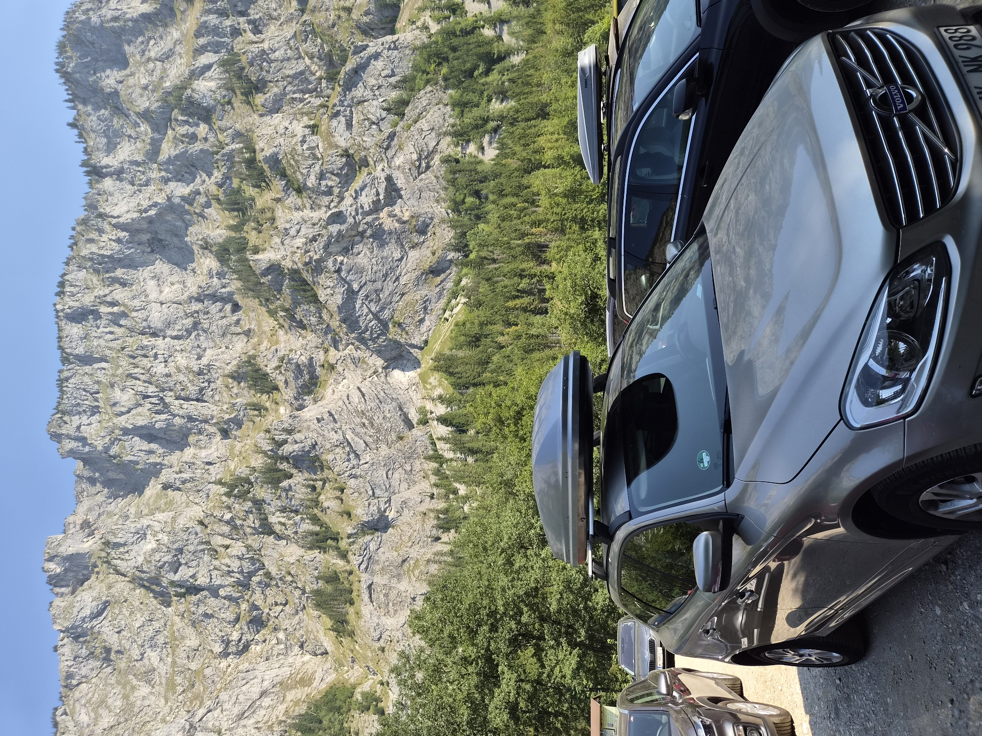 A panoramic view of a parking lot at the base of a steep, rocky mountain face in Eisenerz, Austria. Several vehicles, including a silver Volvo SUV with a cargo box on its roof, a dark SUV with a cargo box, and a gray hatchback, are parked on a gravel surface. Dense green trees line the base of the mountain, which is covered in jagged rock formations and sparse vegetation. The sky is clear and blue.