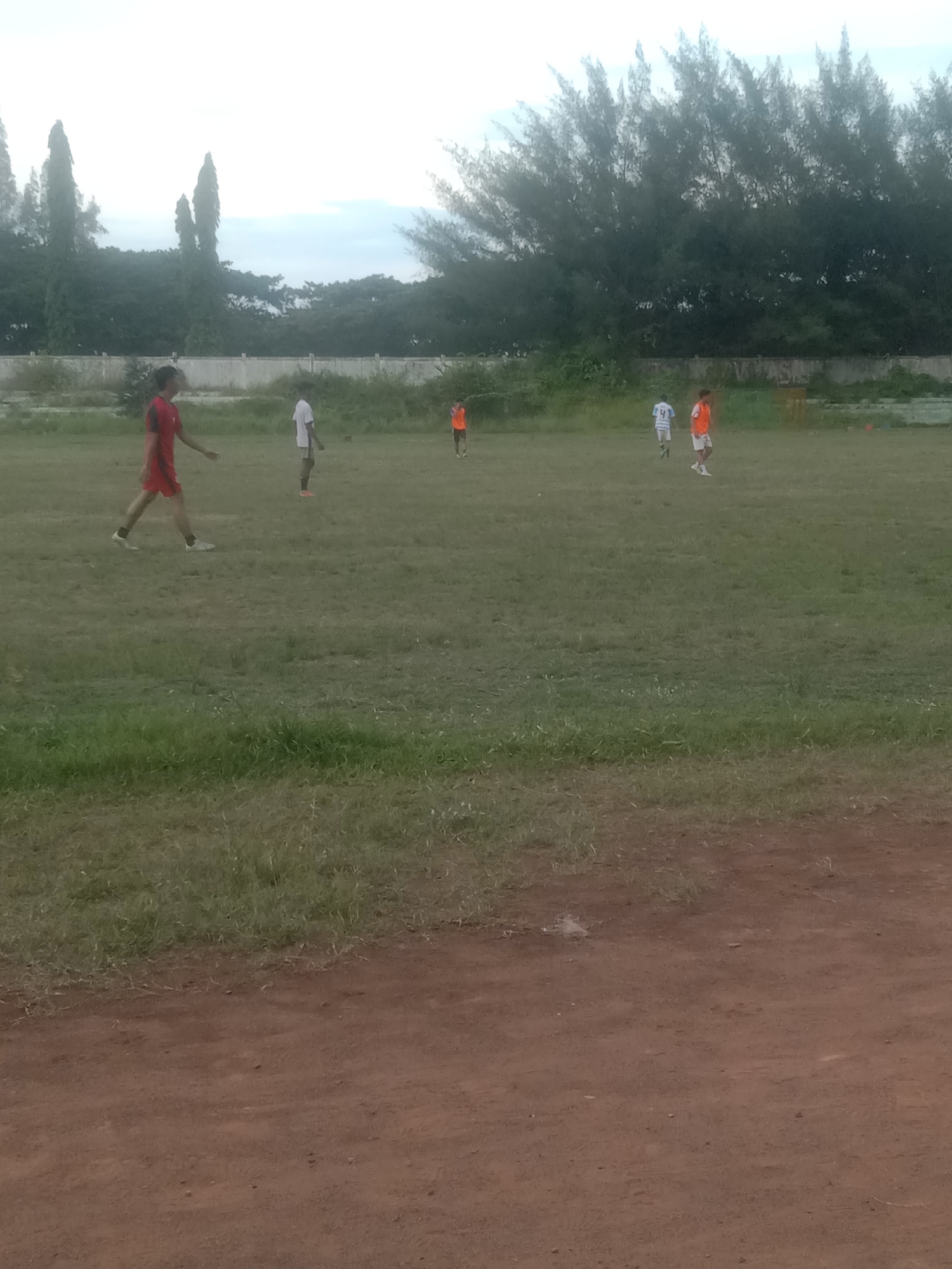 The image captures a soccer field scene in Lhokseumawe, Indonesia, under an overcast sky. The field is grassy with patches of exposed earth, particularly in the foreground. Five individuals are scattered across the field, dressed in athletic attire. One is in a red uniform, another in white, and two in orange jerseys. A white wall spans the background, lined with tall trees. The foreground consists of reddish-brown dirt.