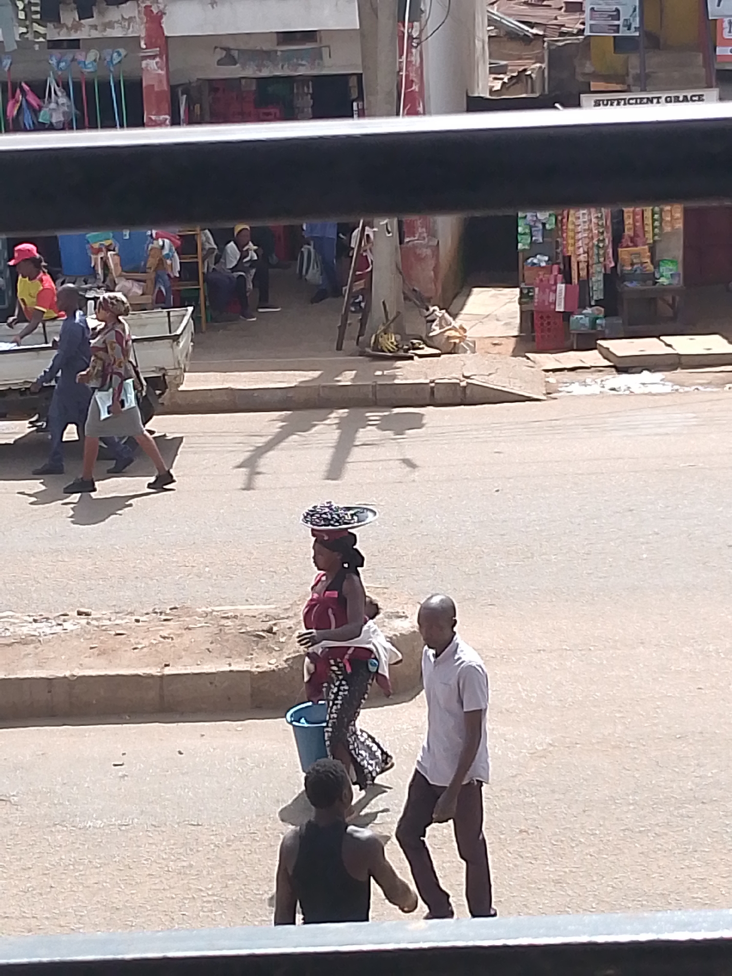 A medium shot captures a street scene in Bukuru, Nigeria, on a bright, sunny day. In the foreground, a dark horizontal bar obscures the bottom of the frame, suggesting the vantage point is from behind a window or barrier. A man in a black tank top stands facing away from the camera, partially visible. To his left, a woman in a maroon and white patterned dress walks, carrying a large tray of items on her head and a blue bucket in her left hand. To her left, a man in a light pink collared shirt and dark pants walks forward. Further left, a woman in a colorful patterned dress walks alongside a man in a dark blue outfit and a white shirt. They are walking past a white truck. In the background, several stalls display various goods, including colorful bags and packaged items. A sign reading "SUFFICIENT GRACE" is visible on a building. The ground is a mix of dirt and asphalt.