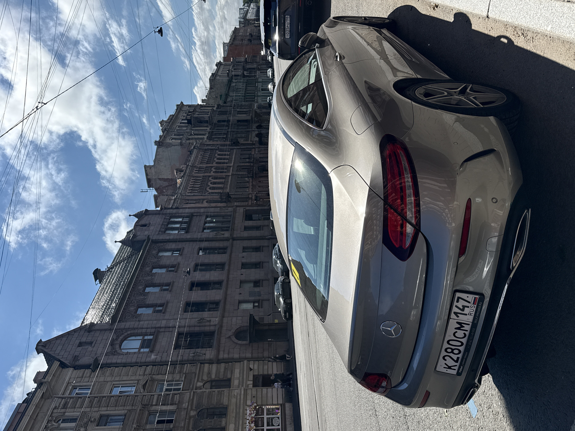 The image shows a parked Mercedes-Benz C-Class coupe on a street in Saint Petersburg, Russia. The car is silver and faces away from the camera, showing its rear. The license plate is visible and reads "K280CM 147". In the background, there are several multi-story buildings with ornate architecture typical of Saint Petersburg. The sky is blue with some white clouds. A dark-colored SUV is parked behind the Mercedes. Overhead, there are visible power lines and streetlights. The scene appears to be a sunny day.