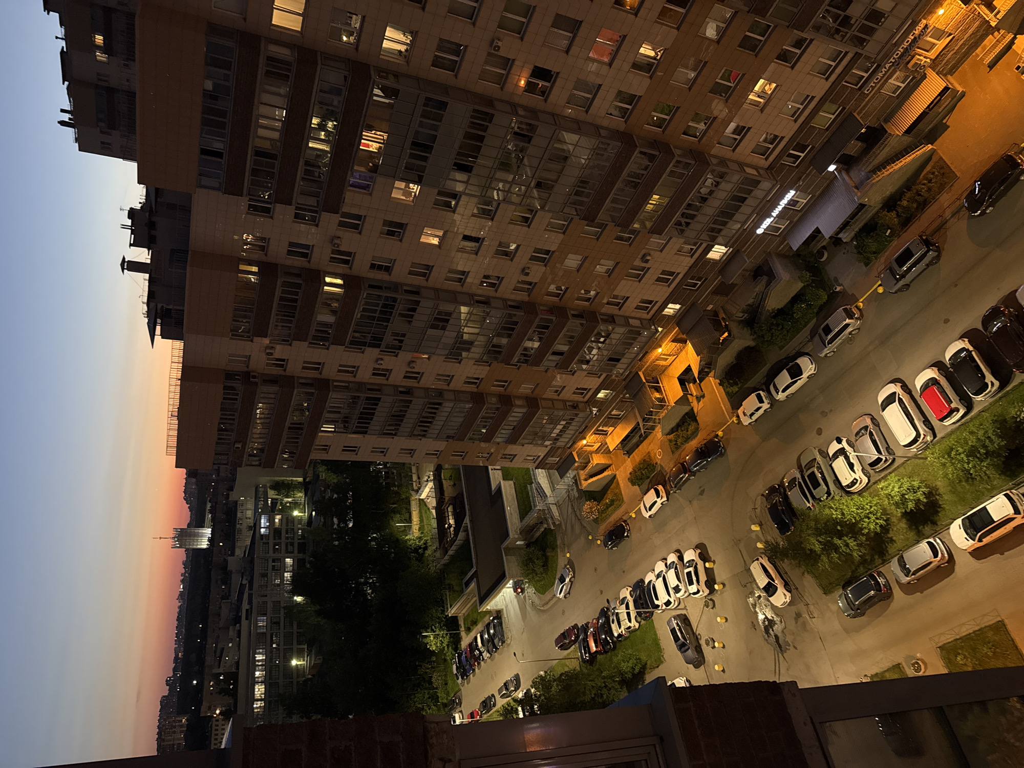 The image captures a dusk scene in an urban residential area, likely in Unknown, Russia. A tall, modern apartment building dominates the right side of the frame, its windows illuminated with warm interior lights, suggesting people are home. Across the street, another apartment building is visible, also lit from within. In the distance, a skyline of other buildings and a distinctive tall tower with a spire can be seen against a gradient sky of orange, pink, and blue. A parking lot in front of the main apartment building is filled with numerous cars of various makes and models. Some cars are parked in designated spots, while others are scattered. A few streetlights cast pools of yellow light onto the asphalt and surrounding green spaces. The overall impression is a quiet evening in a densely populated urban environment.