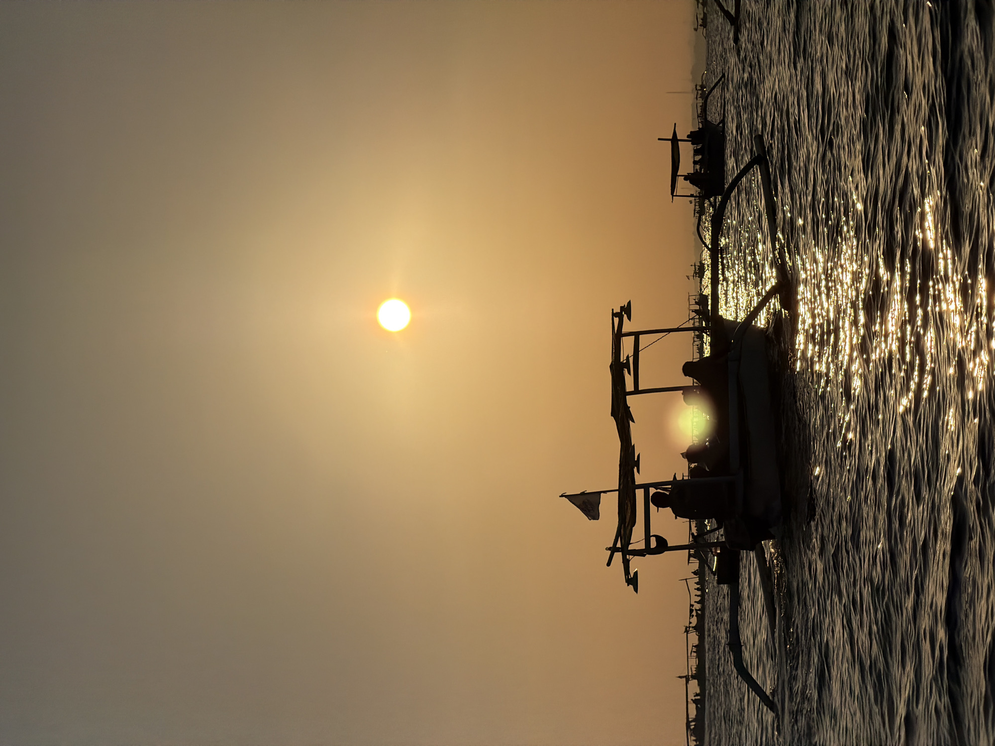 The image depicts a sunset over the sea in Buleleng, Indonesia. In the foreground, a traditional Balinese fishing boat, known as a jukung, is silhouetted against the water. The boat has outriggers extending from its sides, providing stability. Several people are visible sitting in the boat, also in silhouette. A flag is flying from a pole on the boat. The sun, a bright disc, is visible in the upper right portion of the sky, casting a shimmering reflection on the water. In the background, other fishing boats and a hazy shoreline are visible. The sky is a gradient of warm orange and yellow hues, characteristic of a sunset. The sea is choppy, with small waves reflecting the sunlight.