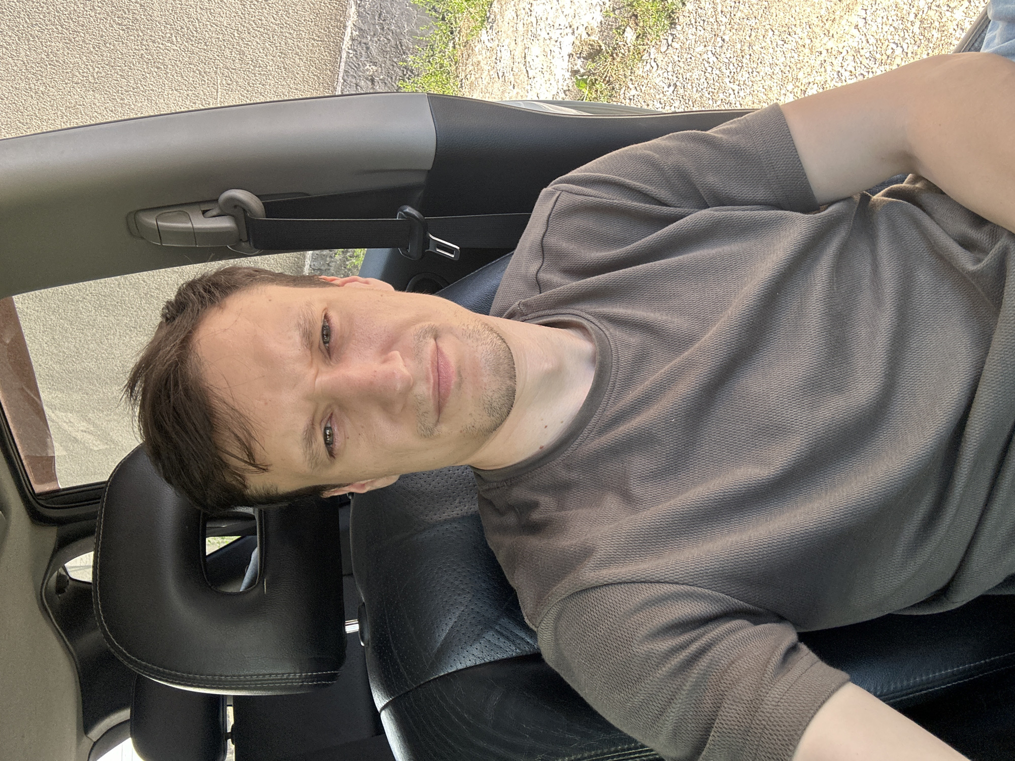 A man is sitting in the driver's seat of a car, facing the camera. He is wearing a brown t-shirt and has short brown hair and stubble. The car's interior is visible, including black leather seats and a headrest with a cutout. The seatbelt is buckled. Outside the car, a textured wall and gravel ground are visible. The scene appears to be in Shatava, Ukraine, on a sunny day.
