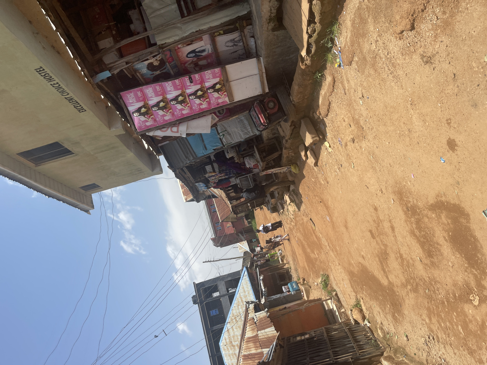 This media file captures a street scene in Jos, Nigeria, likely during daylight hours. The image is oriented vertically, presenting a view looking down a dusty, unpaved street lined with small shops and buildings. The foreground is dominated by the dirt ground, with scattered debris and some patches of dry grass.
On the left side of the street, a building with the text "EXCELLENT CHOICE HOSTEL" prominently displayed on its facade stands out. Adjacent to it are several market stalls, some covered with corrugated metal roofing. These stalls display various goods, with vibrant pink posters featuring images of women and text such as "LUSH," "BE CHIC," and "BUY HERE" being particularly noticeable. These suggest the sale of beauty products or fashion items.
Further down the street, more buildings can be seen, some appearing to be under construction with exposed concrete and metal frameworks. Others are older, constructed from corrugated iron sheets and brick. A few individuals are visible in the distance, engaged in what appears to be daily activities on the street, though their specific actions are not clear. Overhead, a network of electrical wires crisscrosses the sky, indicating the presence of electricity in the area. The sky is partly cloudy, suggesting fair weather. The overall impression is of a bustling, somewhat informal commercial area in a Nigerian city.