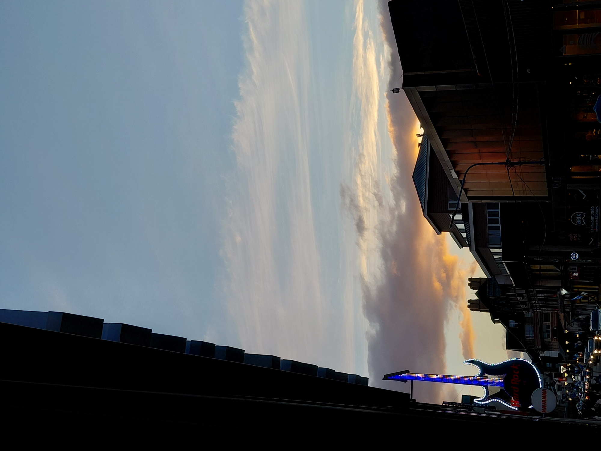 The image shows a street scene in Ushuaia, Argentina, at dusk or dawn. The sky is a mix of pale blue and orange clouds. On the right side of the image, buildings line the street. A Hard Rock Cafe sign, shaped like a guitar with blue and white lights, is visible on the street. People and vehicles can be seen in the background of the street. In the foreground, the dark silhouette of a rooftop is visible.