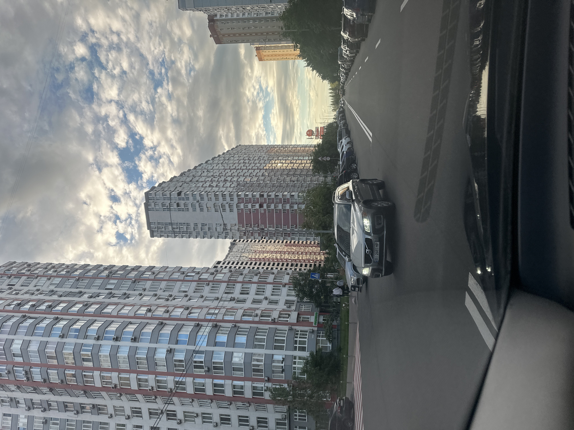 The image depicts a street scene in Kyiv, Ukraine, characterized by modern high-rise buildings and a roadway with several vehicles. Tall, multi-story residential buildings with predominantly white facades and red accents dominate the left and right sides of the frame. The buildings exhibit numerous windows and balconies, indicative of an urban residential area. A grey SUV is prominently positioned in the foreground, driving on the right side of the road. Other cars are parked along the sides of the street and are also visible in motion further down the road. The sky above is partly cloudy, with a mix of white and grey clouds suggesting either early morning or late afternoon light. The overall impression is one of a typical day in a developed urban environment.