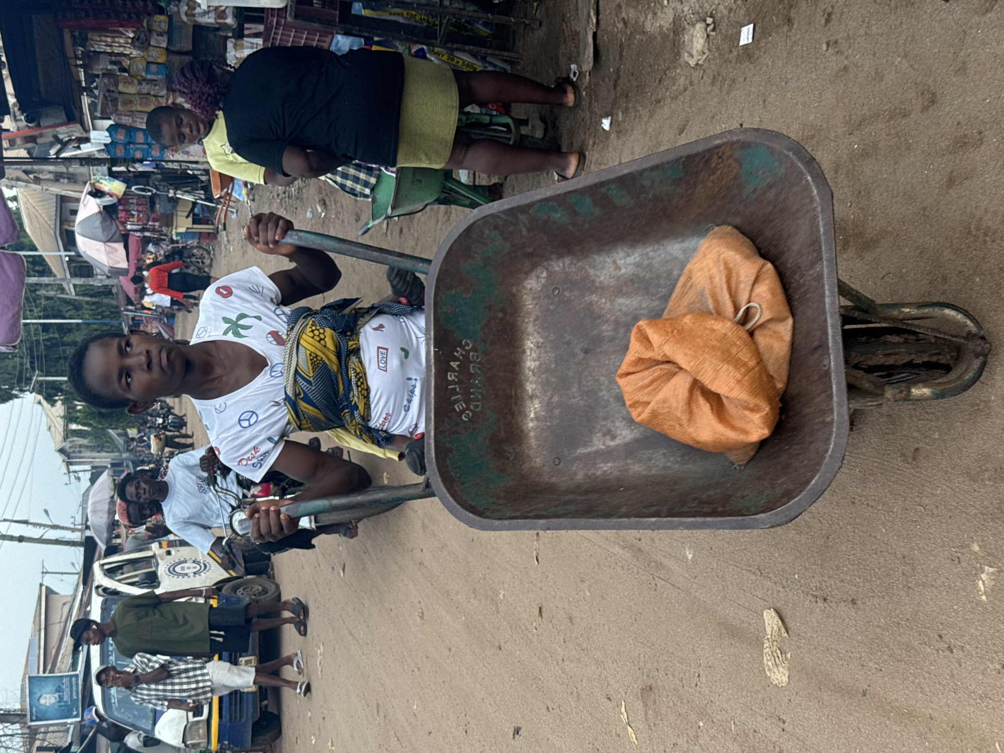 This photograph captures a bustling street or market scene in Ihiagwa, Nigeria, under an overcast sky during the daytime. The image is presented vertically, but depicts a horizontally unfolding scene of daily life and commerce.
In the immediate foreground, a young person with dark skin and short dark hair is pushing a well-used, metallic wheelbarrow. She is wearing a white t-shirt featuring symbols like a palm tree and a peace sign, and the word "LOVE." A colorful, patterned fabric is wrapped around her lower torso. She looks directly at the camera with a neutral expression, holding the wheelbarrow's handles.
The wheelbarrow itself is a prominent feature, showing signs of wear with rust and a greenish-brown patina. It contains two loosely packed, orange-brown sacks, likely holding goods. Faint, mirrored text is visible on the wheelbarrow's side, appearing to read "CHARLIE," possibly "BROTHERS," and numbers like "2007."
Behind the central figure, the street is populated with various individuals. To her right, another person is partially visible, dressed in a black top and a light-colored skirt. Further back, more people are seen, including one in a yellow shirt. To the left, a group of men stands near a blue and white commercial vehicle, which displays a poster on its side. A tricycle, commonly known as a keke, is also visible in the mid-ground.
The background shows numerous market stalls lined with various packaged goods, including what appears to be biscuits (with text "20S FOR ANNA" visible on one package). Several open umbrellas provide shade over the stalls. The ground is a mix of dirt and compacted earth, typical of an outdoor market environment. The scene depicts a vibrant hub of community activity and trade.