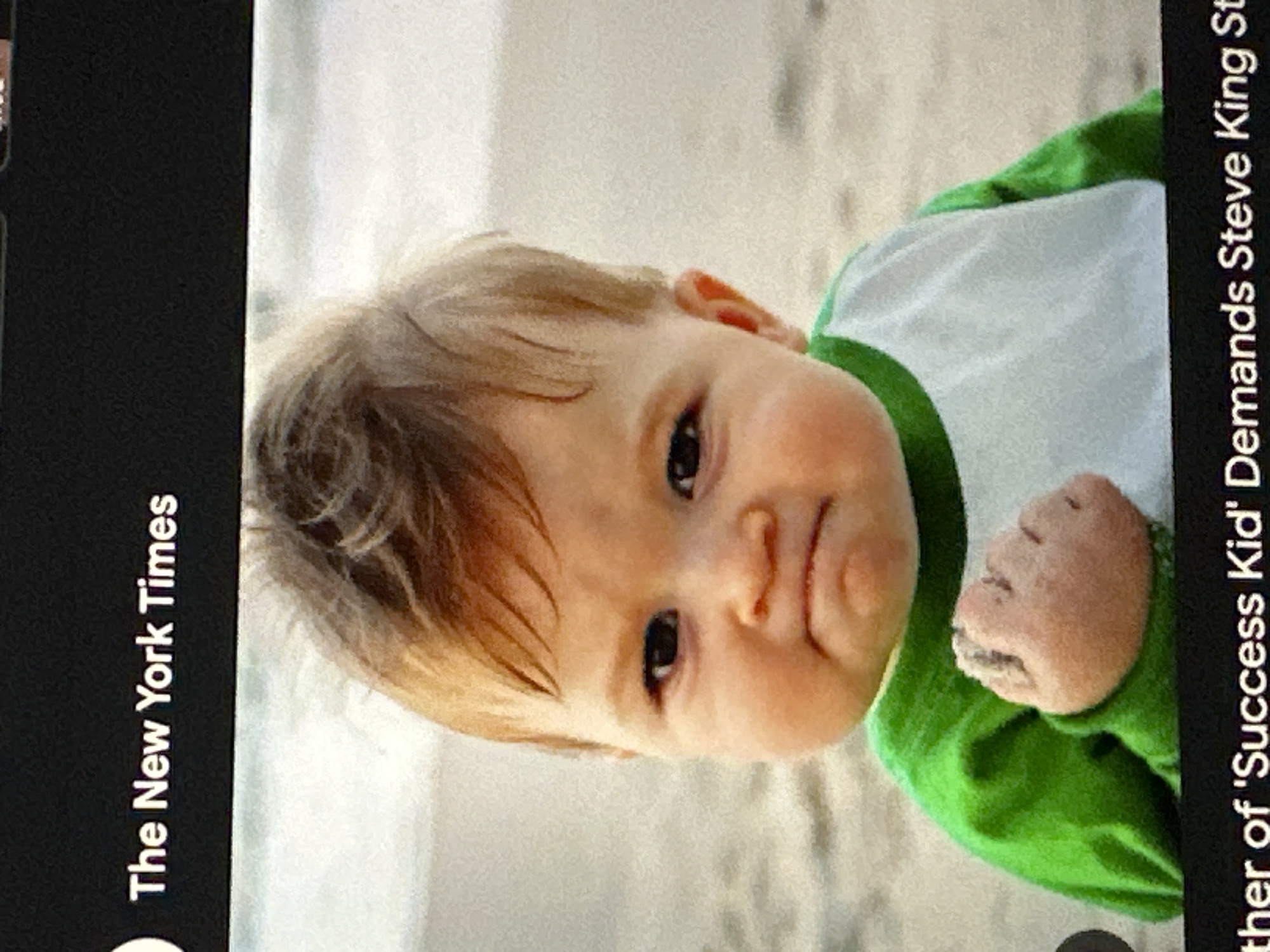 The image displays a screenshot of a digital screen featuring a news article attributed to "The New York Times". The primary subject is a light-skinned infant with light brown hair, dark eyes, and a determined or defiant facial expression characterized by furrowed brows and a slightly downturned mouth. The infant wears a green long-sleeved shirt with a white front panel. The right hand is clenched into a fist, with light-colored sand particles visible on the knuckles. The background consists of a sandy beach, with a subtle horizon line in the distance. This beach scene is located in the city of Ta Xua, Vietnam. A partial headline at the bottom of the article reads, "ther of 'Success Kid' Demands Steve King St".