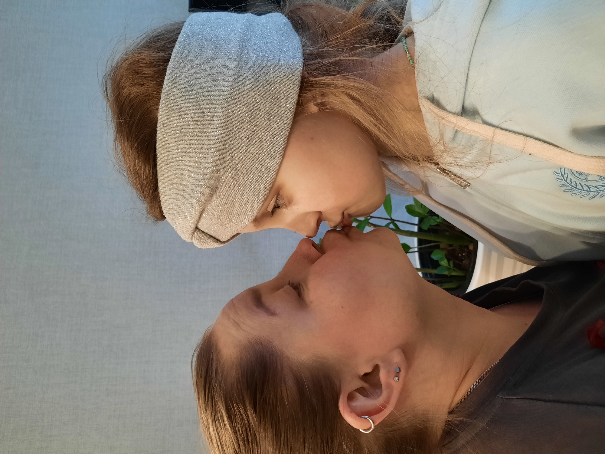Two individuals, one appearing younger, are positioned close together, with their faces touching. The younger individual wears a light-colored headband and a light blue hooded sweatshirt with a delicate necklace visible at their neck. The other individual has multiple piercings in their ear and is wearing a dark-colored shirt. A potted plant is visible in the background. The location is not identifiable as the city of Pervomaysky, Russia.