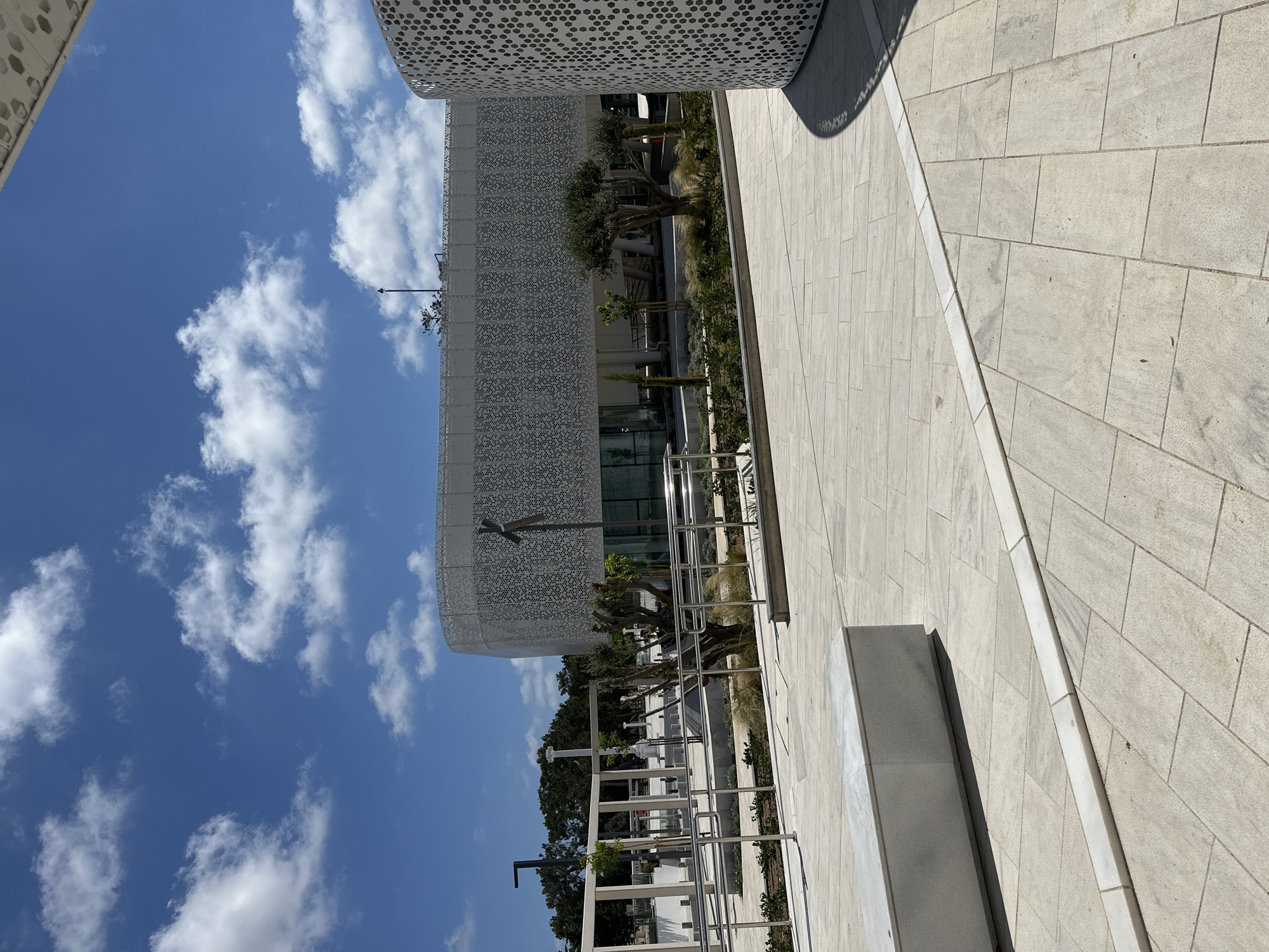 The image depicts the exterior of the new Municipal Market building in Paphos, Cyprus, under a clear blue sky with scattered white clouds. The building features a modern, curved facade with a white perforated screen. In the foreground, a paved plaza area is visible, laid with large, light-colored rectangular tiles. A long, low, grey stone bench sits on the left side of the plaza. A thin white marble strip runs diagonally across the tiled area. Behind the bench, a pathway with metal railings leads towards the building. Landscaping with small trees, shrubs, and dry grass is present between the pathway and the building. The background shows more of the distinctive perforated facade of the Municipal Market and a glimpse of trees and other structures. The scene is static, with no people or animals present, suggesting an outdoor, public space during daytime.