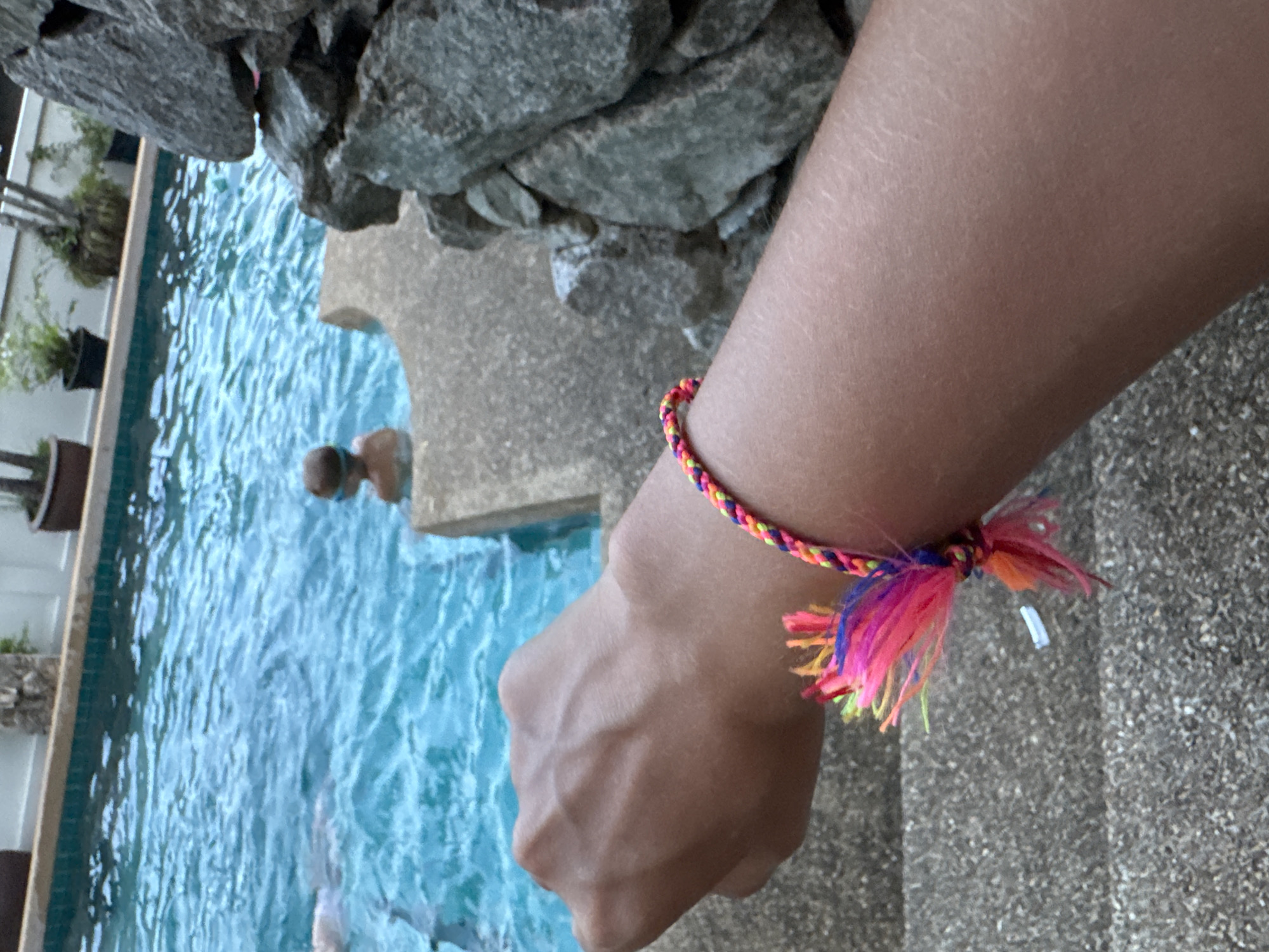 A person is standing next to a swimming pool in Rawai, Thailand. The person is wearing a colorful braided bracelet on their left wrist. The pool has a light blue, rippled surface. Adjacent to the pool are dark gray rocks and potted plants. Another person is visible in the pool, with only their head above water. The area surrounding the person and the pool appears to be paved with textured concrete.