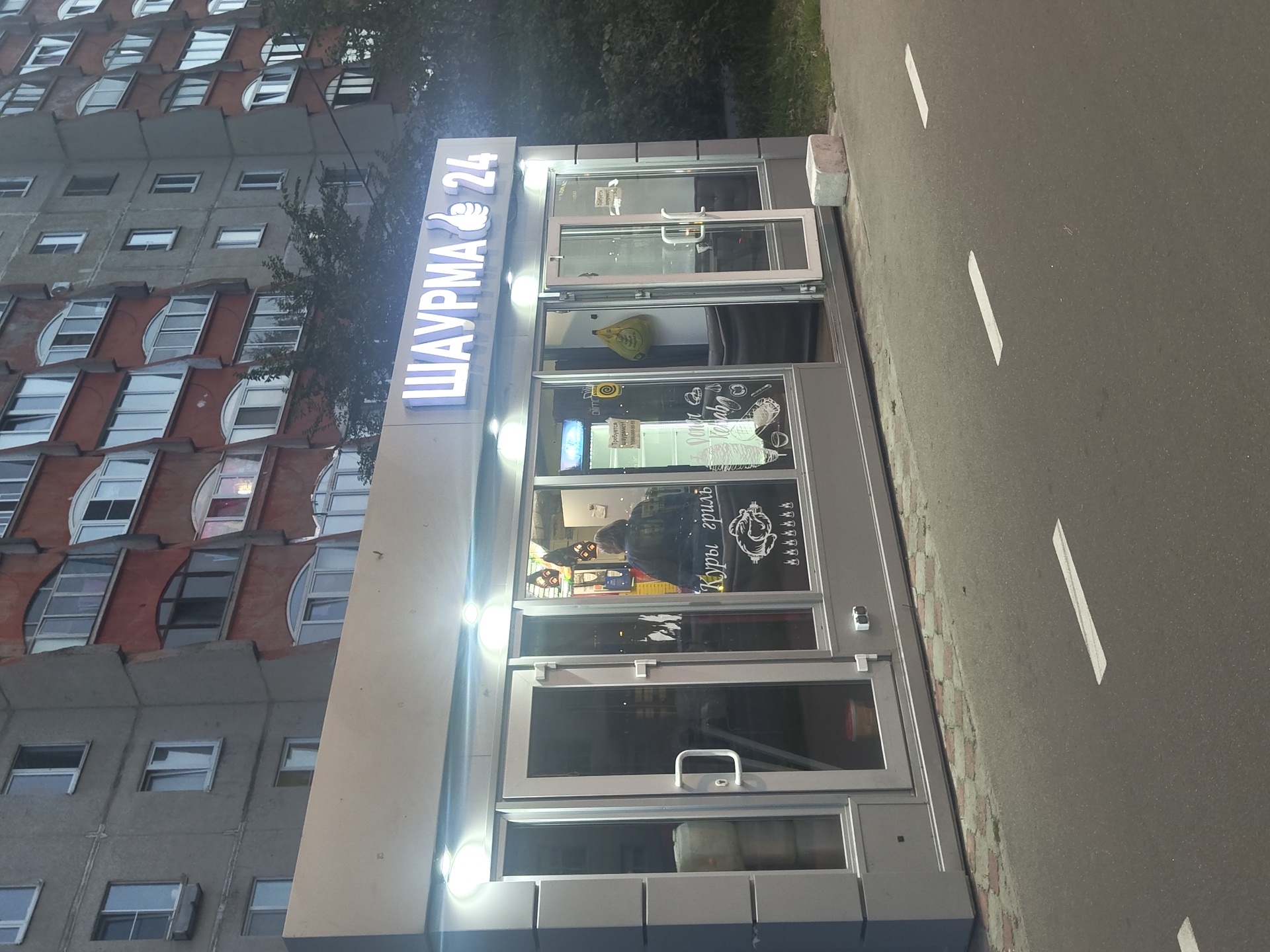 The image shows a street-level view of a food kiosk in Zhukovsky, Russia, at dusk. The kiosk, labeled "ШАУРМА 24" (Shawarma 24) in illuminated white lettering, features a modern grey facade with large glass windows and doors. Inside, a person wearing dark clothing is visible behind the counter, serving customers. The interior displays menu items such as "Куры гриль" (Grilled Chicken) and "Doner Kebab," along with images of food.
The kiosk is situated on a paved street with visible white road markings, suggesting it's a public thoroughfare. Behind the kiosk, a large, multi-story residential building with a distinctive red and grey facade dominates the background. Trees and foliage are visible on the right side of the image, partially obscuring the residential building. A light grey concrete block rests on the sidewalk near the kiosk. The lighting suggests an evening setting, with the kiosk and building illuminated, creating a contrast with the darkening sky.
