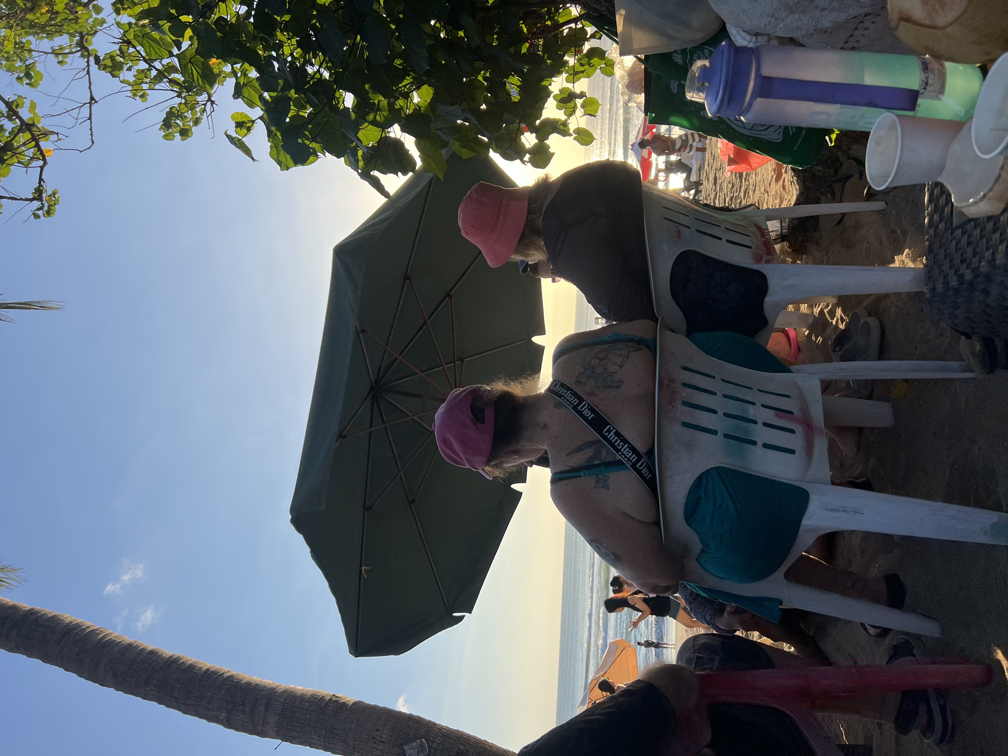 Two people are seated on plastic chairs on a beach in Kuta, Indonesia, facing the ocean. The person on the left, viewed from behind, wears a pink baseball cap and a teal tank top, with tattoos visible on their back. A black and white strap with "Christian Dior" printed on it is worn across their shoulder. The person on the right, also viewed from behind, wears a pink bucket hat and a dark jacket. Both individuals are seated under a large green umbrella, with a palm tree on the left and foliage on the right. In the background, the ocean stretches to the horizon under a bright sky, with other beachgoers visible in the distance. The sandy ground is littered with various objects including cups and a reusable water bottle.