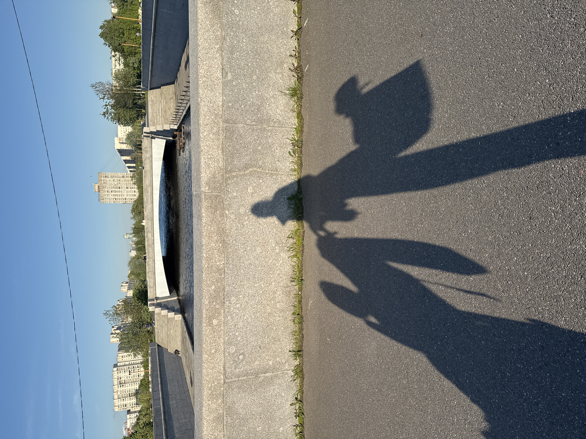 The image depicts a cityscape in Saint Petersburg, Russia, taken from a low vantage point, likely from the ground level. The photo is horizontally oriented with the left side showcasing the cityscape, and the right side displaying a section of pavement where shadows are cast.
The cityscape consists of a river embankment constructed of concrete and granite with several tiers leading down to the river's edge. A bridge-like structure, painted white, arches over the river. Behind the embankment, residential buildings and trees define the urban landscape. The sky is a clear, light blue. A single overhead wire cuts across the upper left portion of the sky.
The right side of the image is dominated by a gray asphalt pavement. The shadows of two individuals are projected onto the asphalt. The outlines of the shadows suggest they are two figures walking, one appearing taller than the other. A thin line of vegetation, consisting of grass and weeds, grows along the edge where the pavement meets a stone curb.