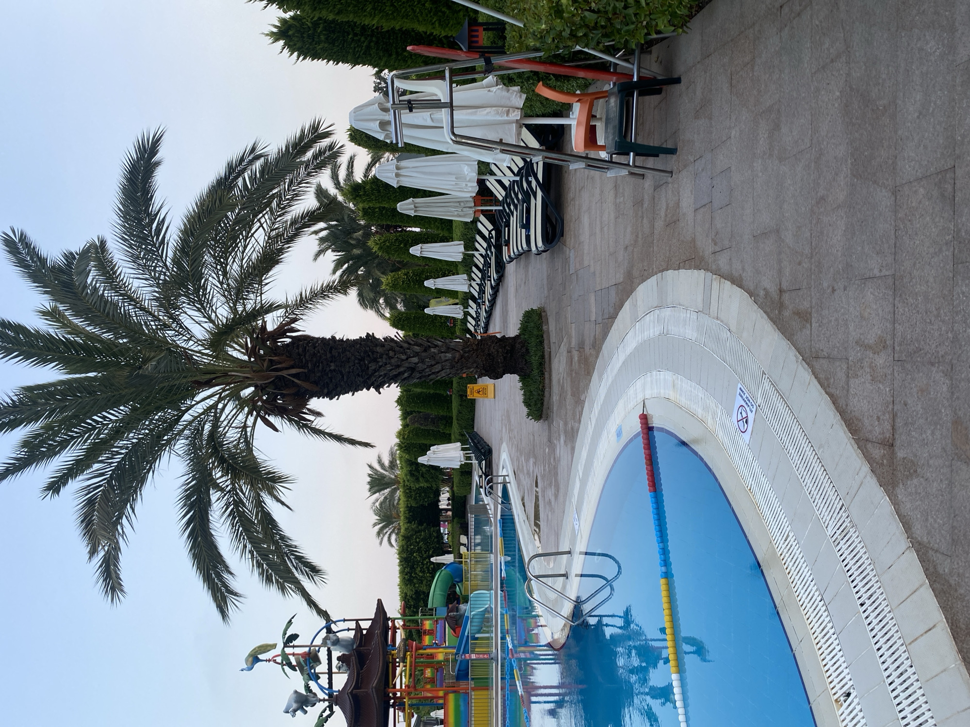 This is a photograph of a swimming pool area at a resort in Aksu, Turkey. The pool is a large, bright blue body of water with lane markers. To the left of the pool is a multi-colored water slide structure with decorative animal figures. A tall palm tree stands prominently in the center of the image. To the right of the pool, a row of lounge chairs and closed white umbrellas are neatly arranged on a paved patio. Adjacent to the lounge chairs is a lifeguard chair with a few chairs stacked at its base. The area is bordered by manicured green hedges and cypress trees. The sky is overcast and the lighting suggests it is either early morning or late evening. There are no people visible in the image.