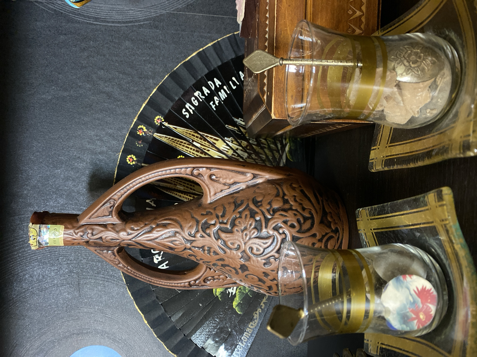 The image depicts a still life arrangement of decorative objects, with no indication of a specific location in Novaya Usman', Russia. In the background, a dark surface, possibly a wall or fabric, serves as a backdrop. A large, brown ceramic bottle with an ornate, embossed pattern and two carved handles stands prominently. Above and behind the bottle, a black fan is partially open, revealing an illustration of the Sagrada Familia. To the right of the bottle, a wooden box with intricate detailing is visible. In the foreground, two clear glass tumblers, each adorned with two gold bands, are placed on decorative glass coasters with gold geometric patterns. Each tumbler contains a small metal spoon and some contents, with the left tumbler holding what appears to be a small, round decorative disc with floral imagery, and the right tumbler containing light brown, granular material, possibly sugar cubes or rock candy. The lighting is subdued, creating shadows and highlighting the textures of the objects. There are no people or actions depicted in the scene.