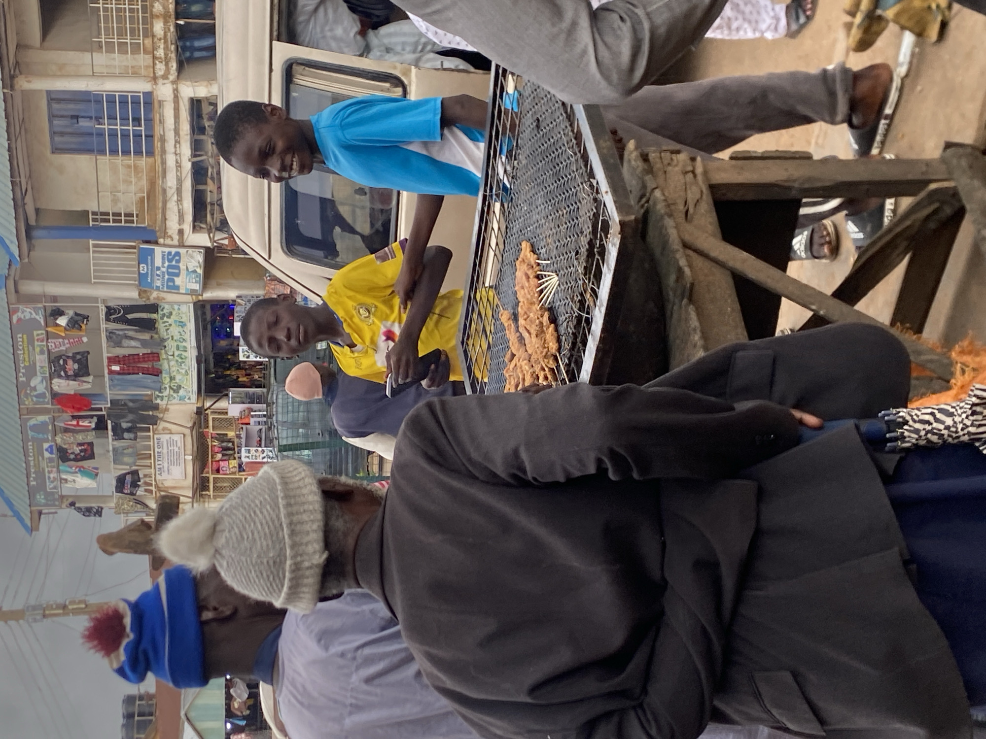 The image depicts a street scene in Bukuru, Nigeria, with a focus on a group of people gathered around a grill. In the foreground, an older person wearing a dark suit jacket and a grey knit hat is facing away from the camera, observing the grill. To their right, two young boys are standing near a beige van. One boy, in a yellow t-shirt, is holding a phone and looking at the camera. The other boy, in a blue t-shirt, is smiling and looking towards the grill.
The grill itself is laden with skewers of meat, presumably being cooked. The grill is supported by a rustic wooden structure. In the background, buildings with storefronts line the street, displaying clothing and various goods. One sign reads "Preston Collection." The overall atmosphere suggests a busy marketplace or roadside food stall. The lighting indicates it is daytime, likely overcast.