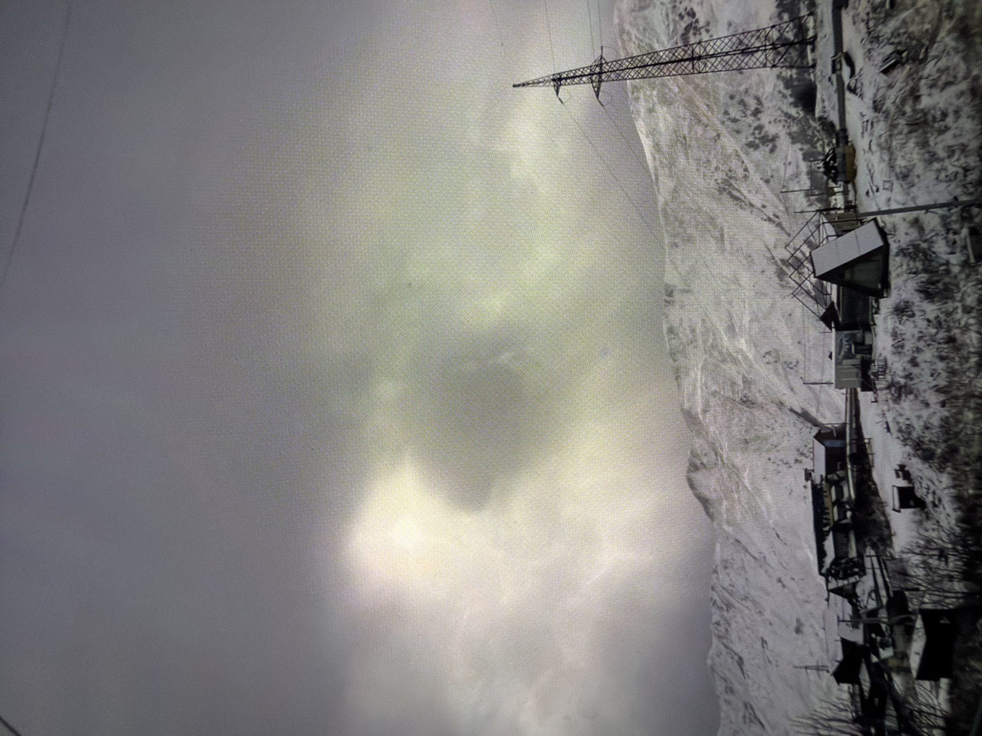 The image shows a snow-covered mountainous landscape under a cloudy sky. A power line tower is visible in the upper-right quadrant of the image. Buildings, possibly residences or industrial structures, are located at the base of the mountain. The location is presumed to be in a city, but the specific city is unknown.