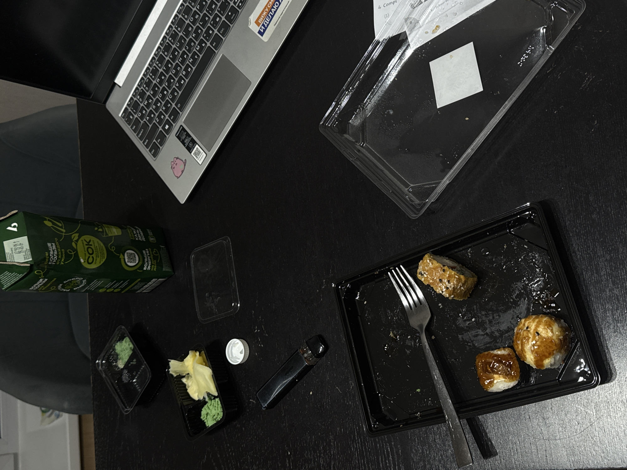 A high-angle shot depicts a cluttered, dark surface. A laptop is partially visible in the upper left corner. Adjacent to it is a translucent, rectangular container, possibly used for takeout food. A green carton, labeled in Cyrillic script and identified as "COK" (juice), sits near two small, open containers filled with wasabi and ginger. A small, white cap is next to a dark, elongated container, likely soy sauce. A black, rectangular tray contains remnants of sushi, along with a fork. Based on the juice carton, the location is likely in Perm, Russia. The scene suggests a meal has been eaten while working on a laptop.
